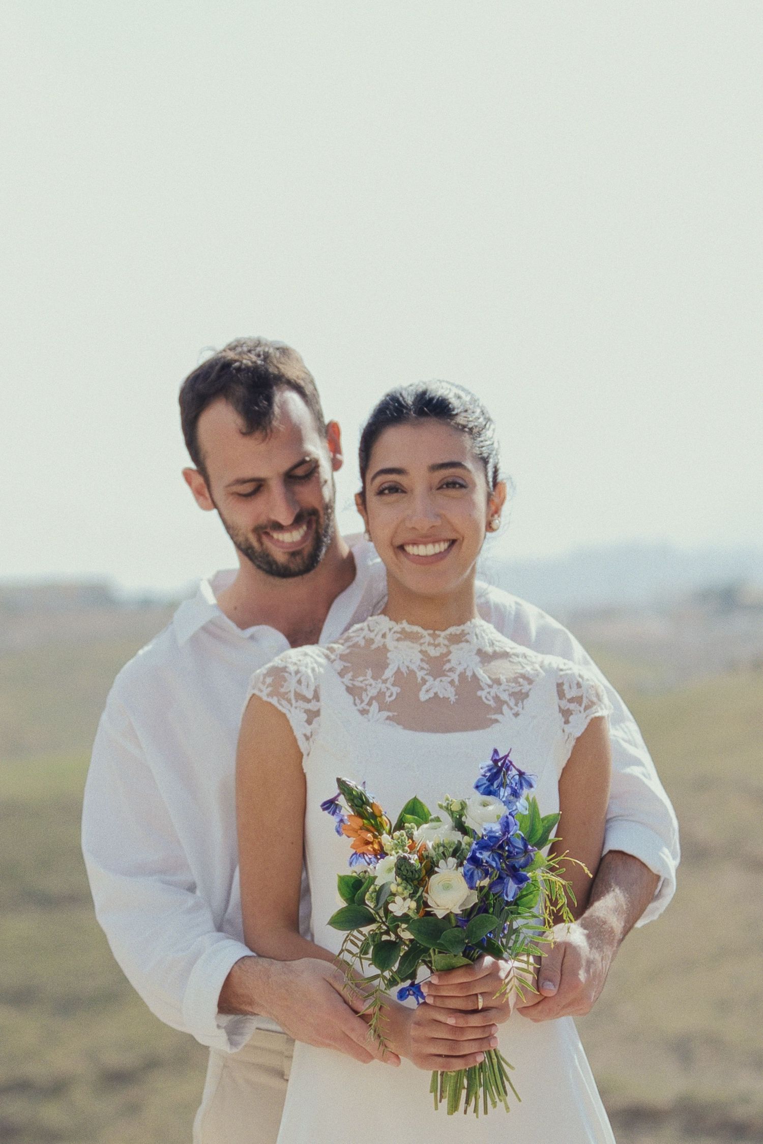A joyful couple embraces in an outdoor setting with the bride holding a colorful bouquet of flowers.