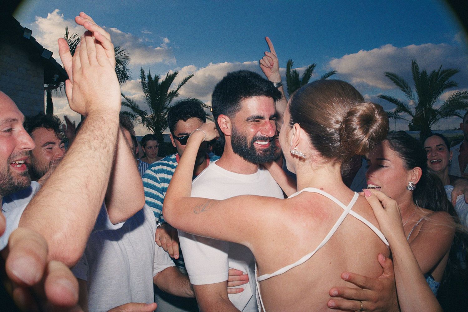 Joyful couple embracing and dancing at an outdoor celebration surrounded by smiling guests with palm trees in background.