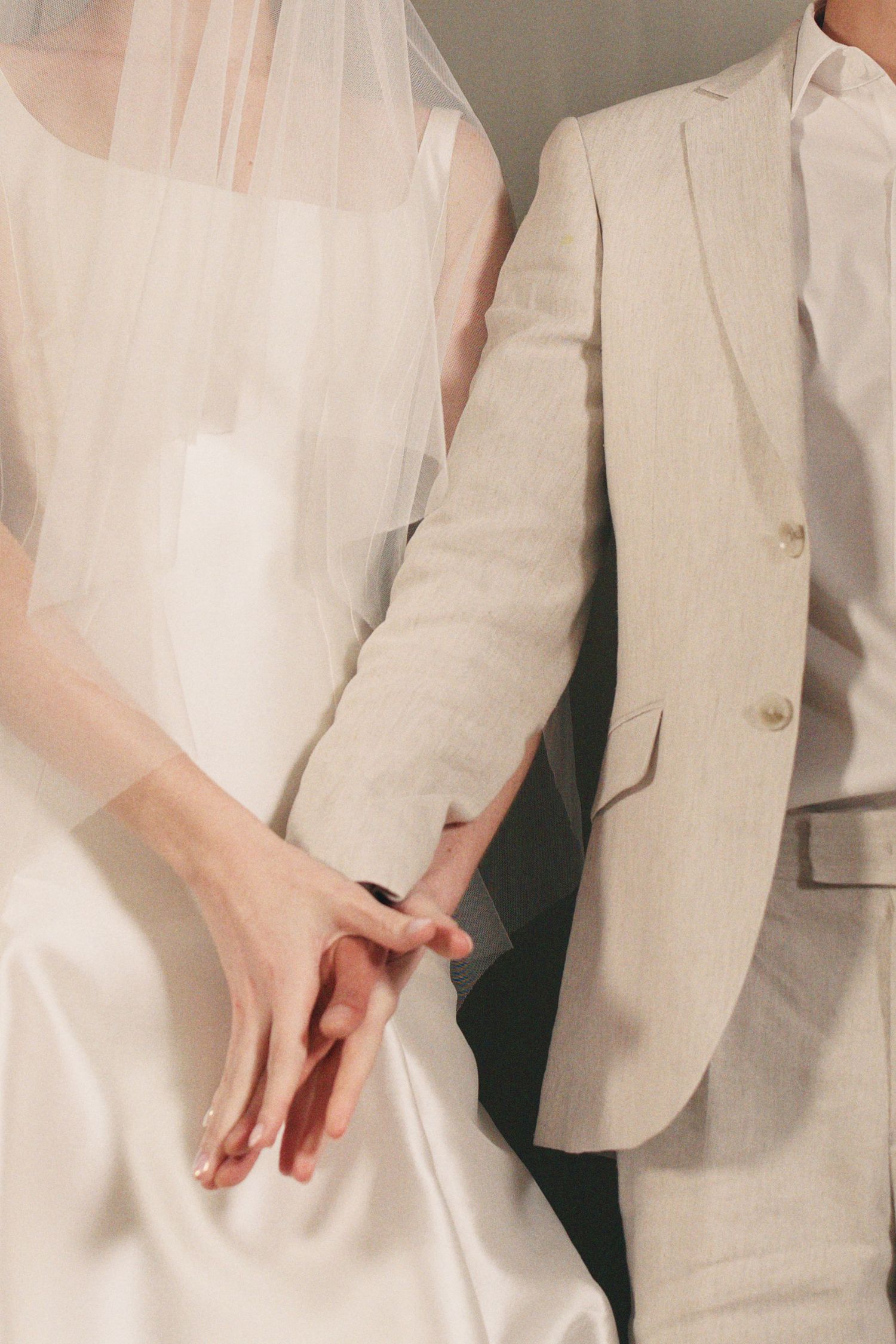 Close-up of bride and groom holding hands, bride in white satin dress and groom in beige suit.