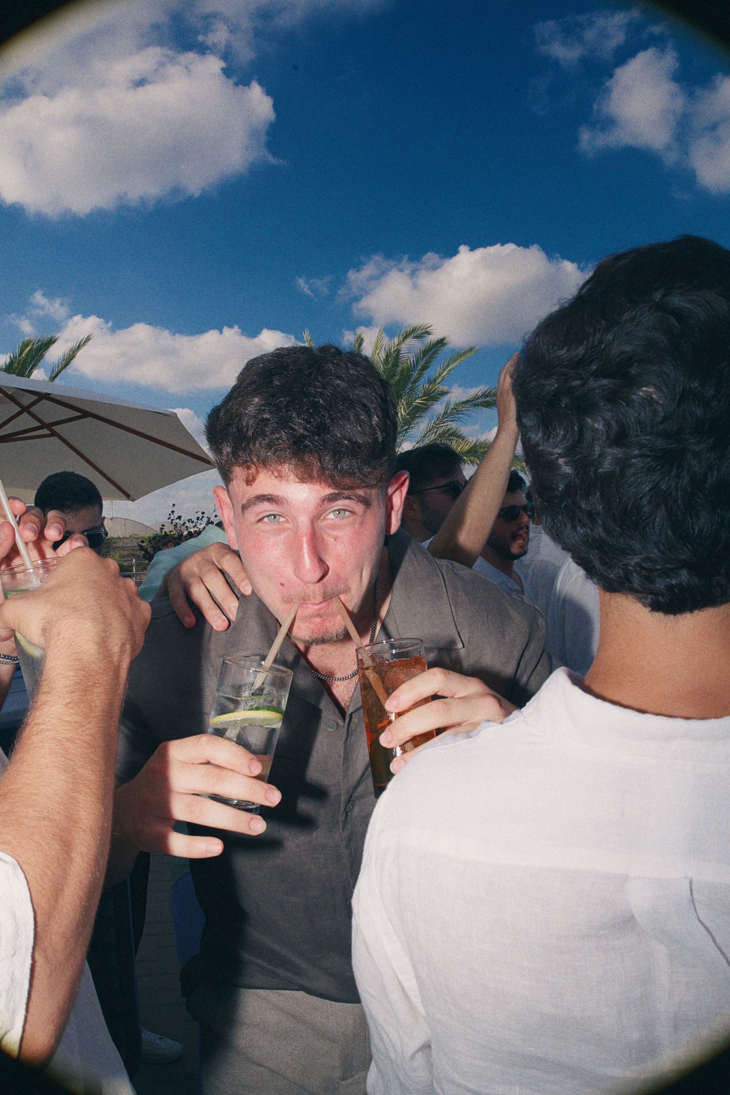 Young man drinking cocktail through straw at outdoor party with friends under blue sky.