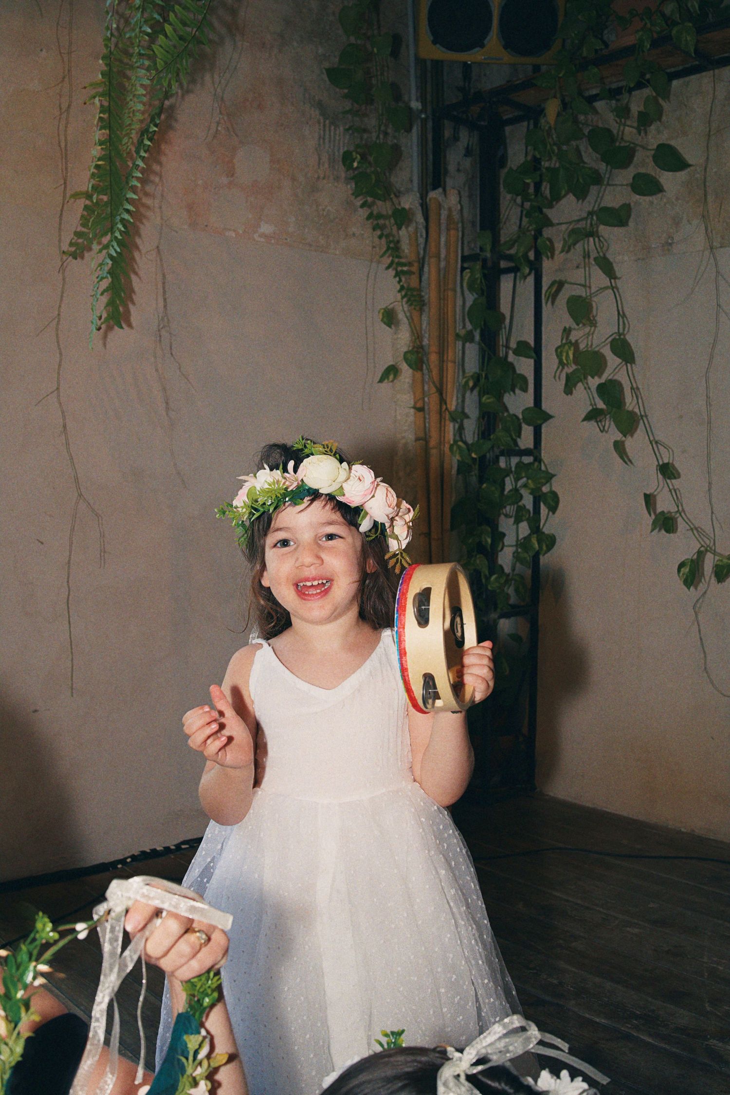 Happy flower girl in white dress and floral crown playfully holds a tambourine at a wedding celebration.