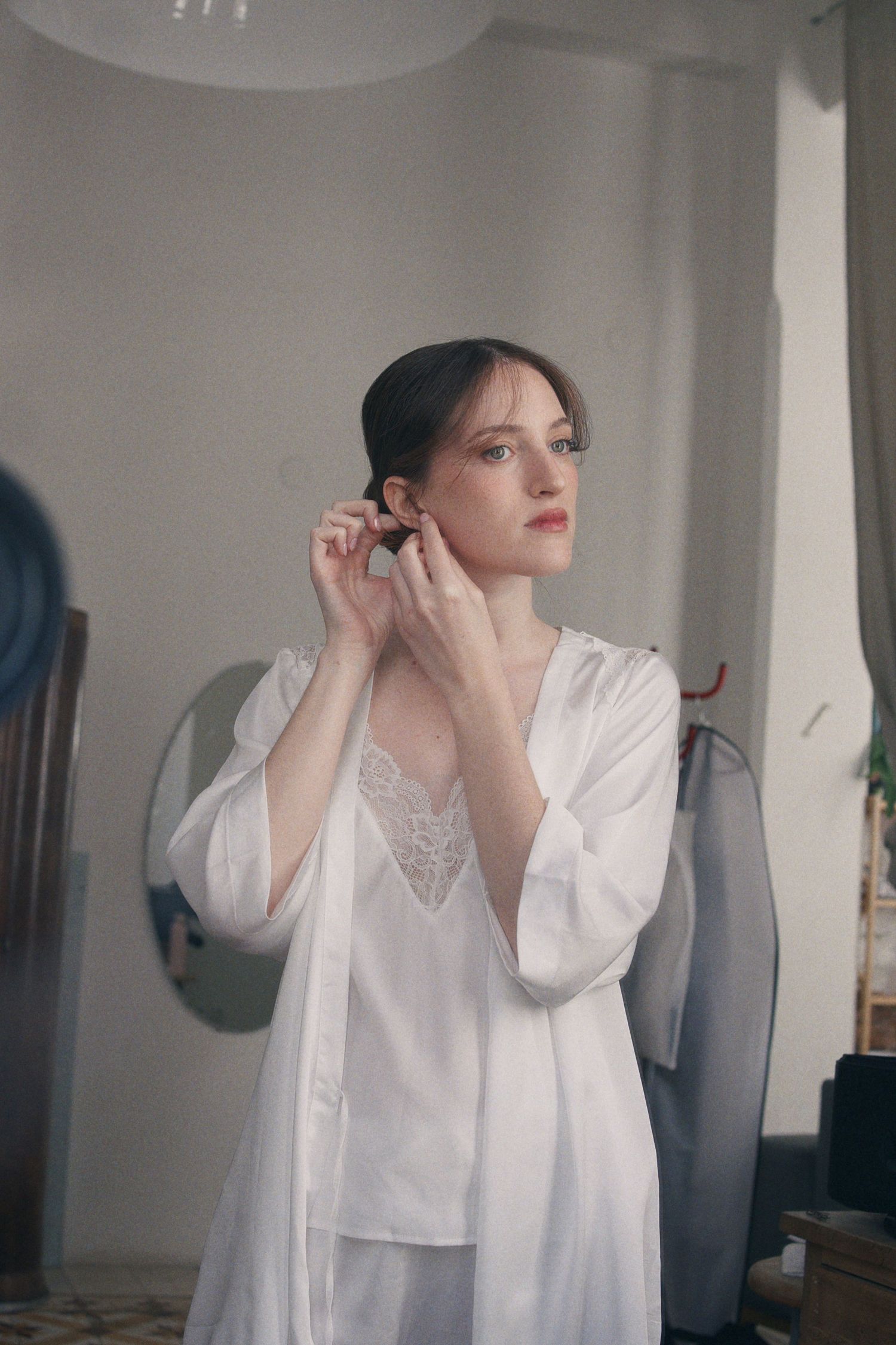 Bride in white silk robe putting on earrings while getting ready on her wedding day.