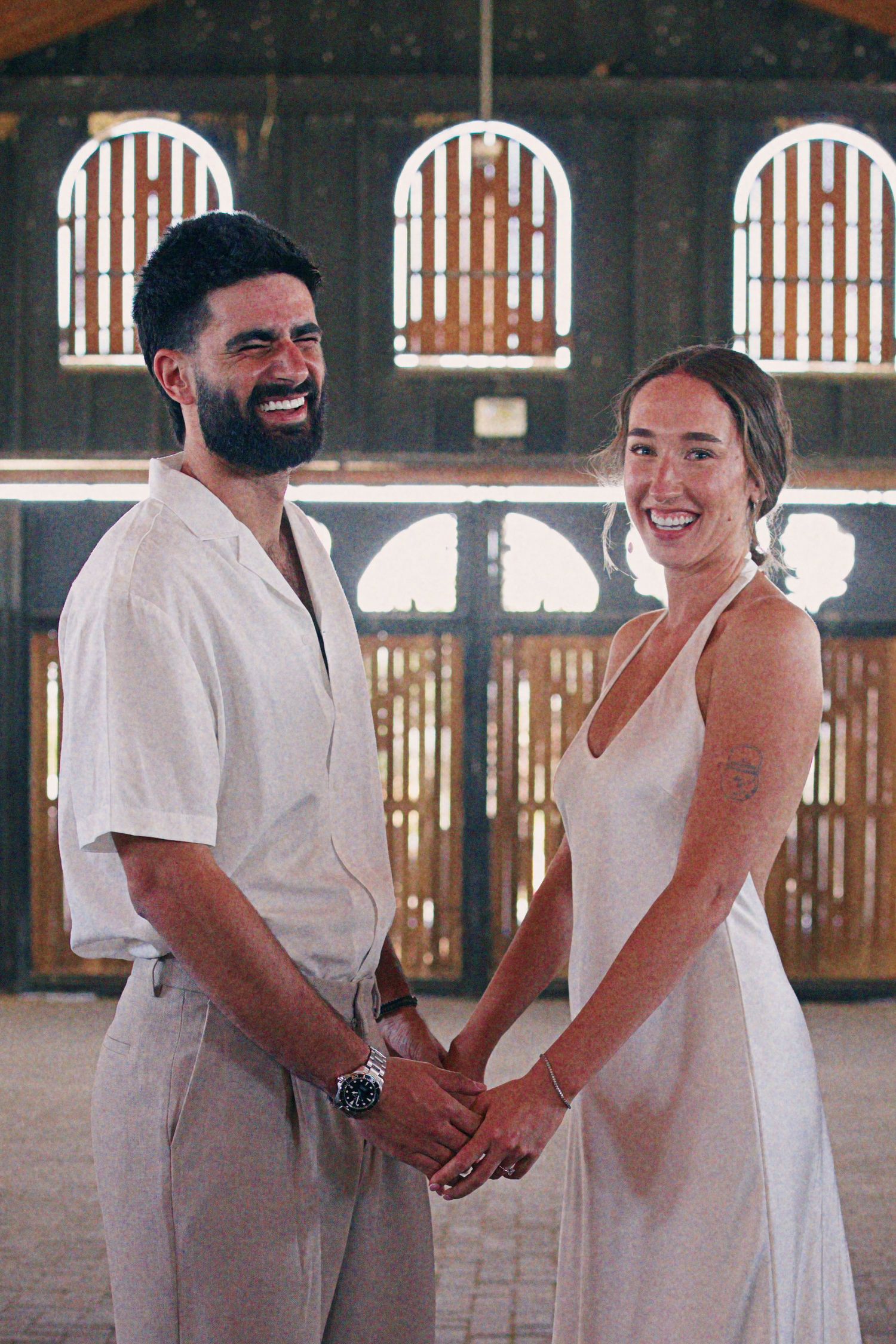 Smiling couple holding hands in white outfits inside a rustic barn venue, celebrating their wedding day.