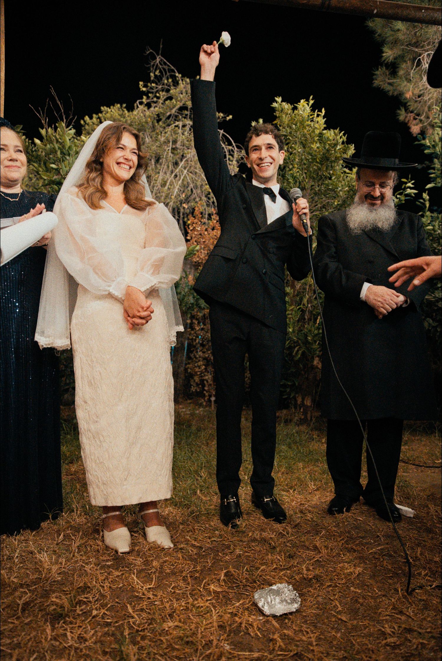 Wedding party members in formal attire pose together on rustic ground during evening celebration.