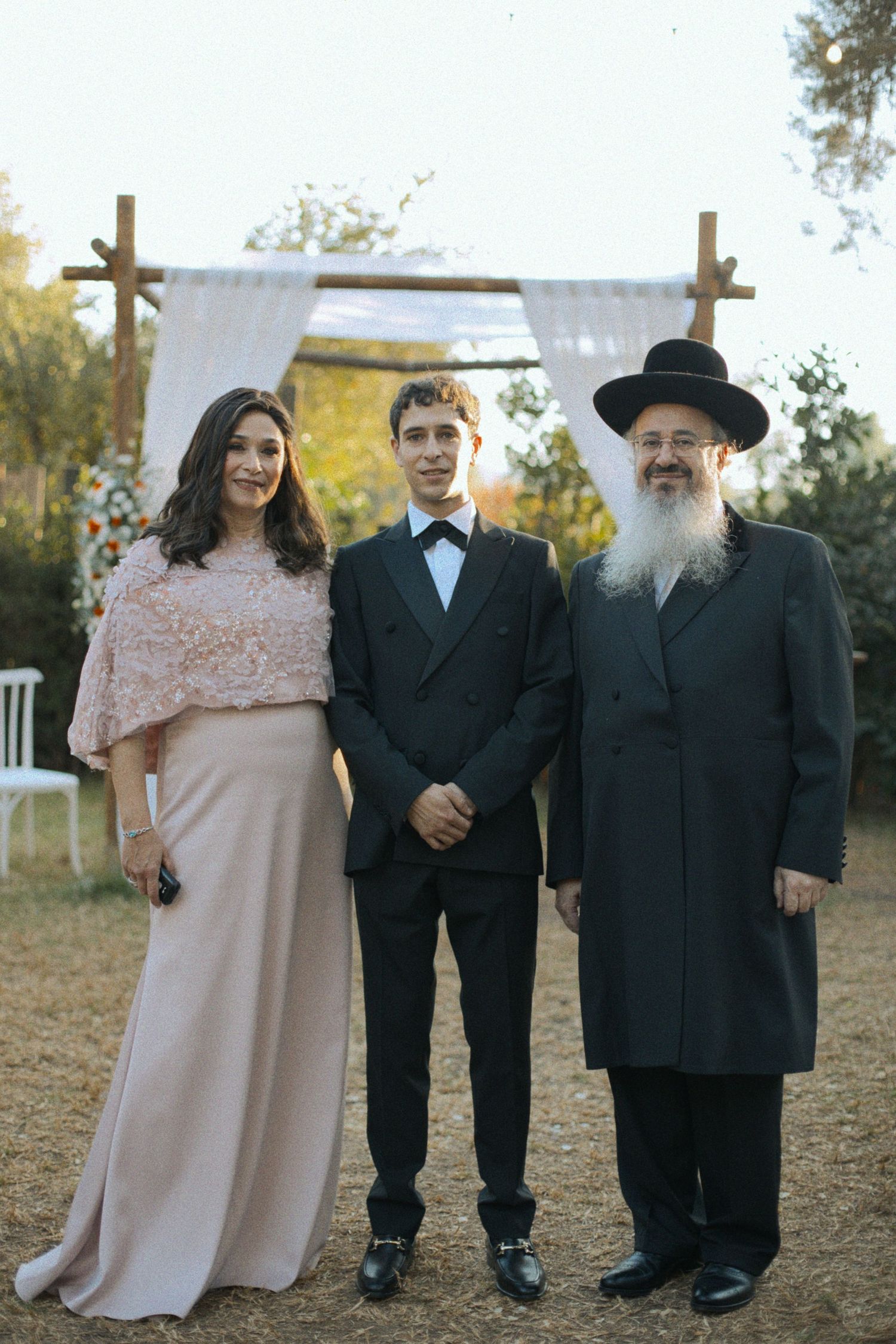 Formal group portrait of wedding attendees in black suits and light colored dresses standing outdoors.