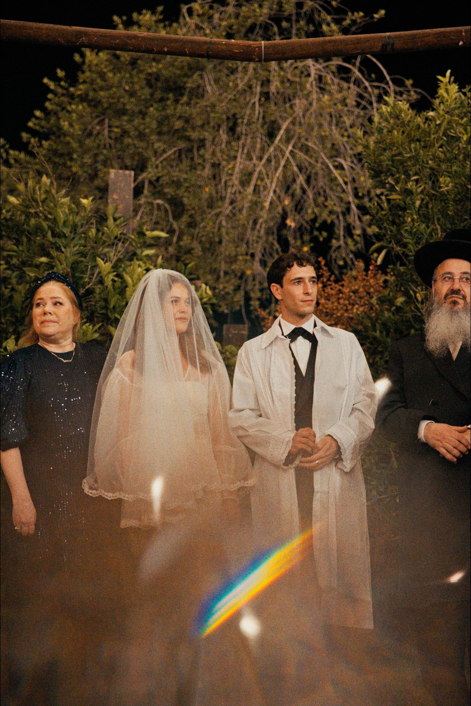 Nighttime wedding ceremony with candlelight creating a rainbow prism effect under wooden arch.