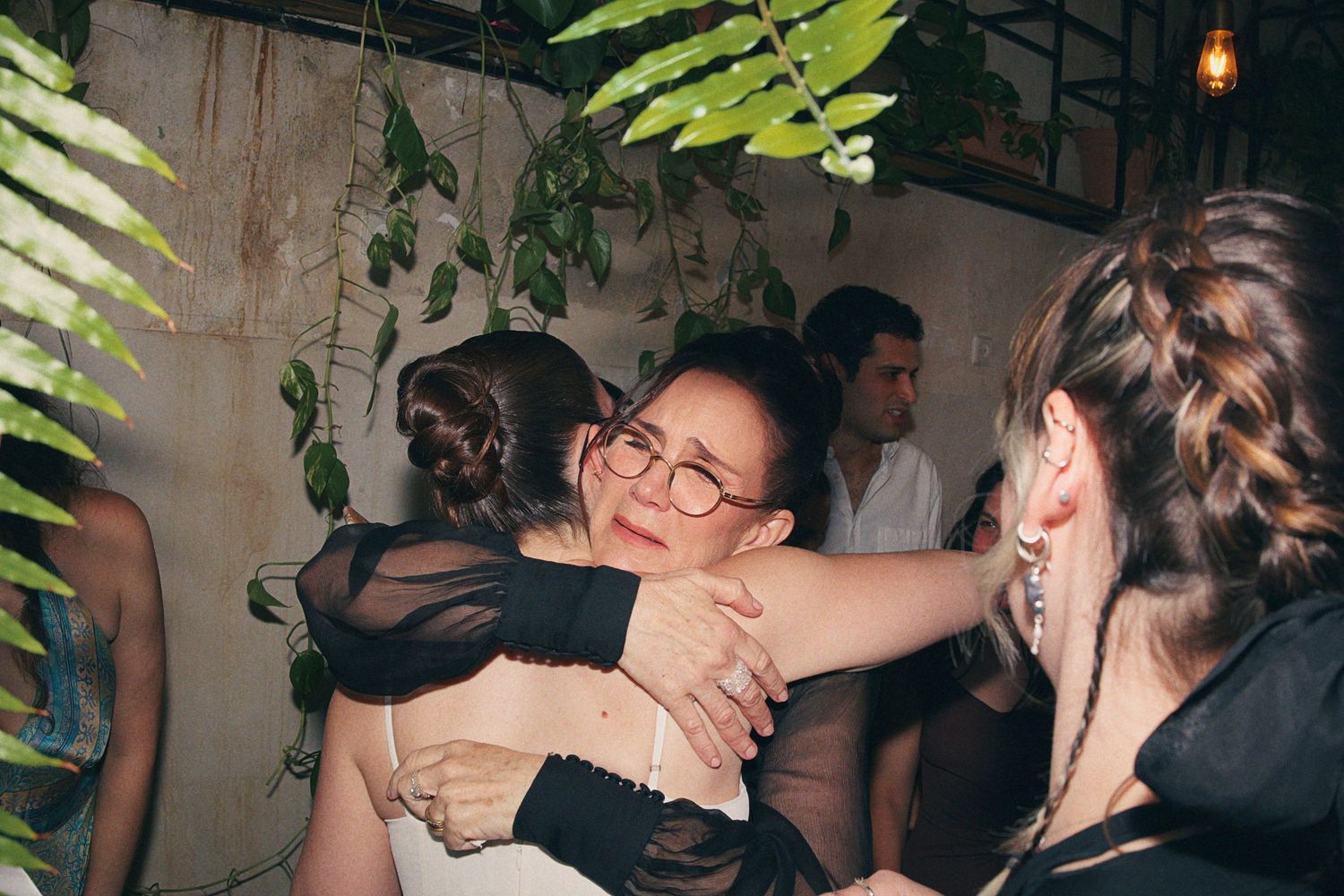 Emotional wedding guest hugging the bride in a white dress at a reception surrounded by greenery.