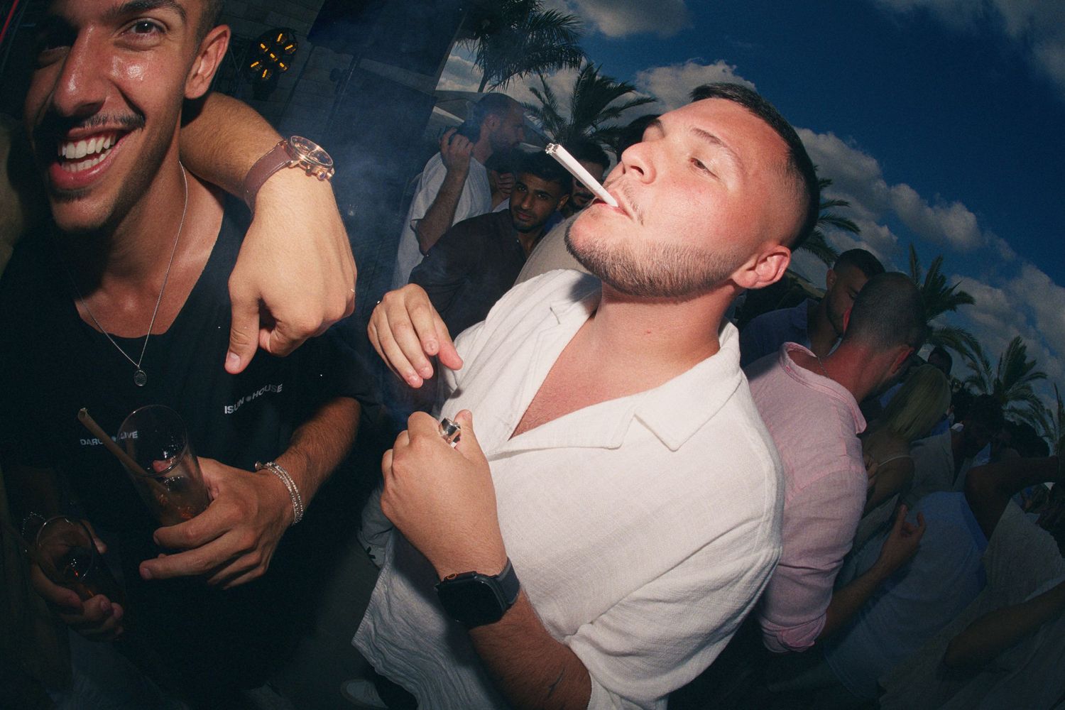 Two men at a party, one smoking a cigarette, posing in front of a tropical backdrop at a nightclub event.