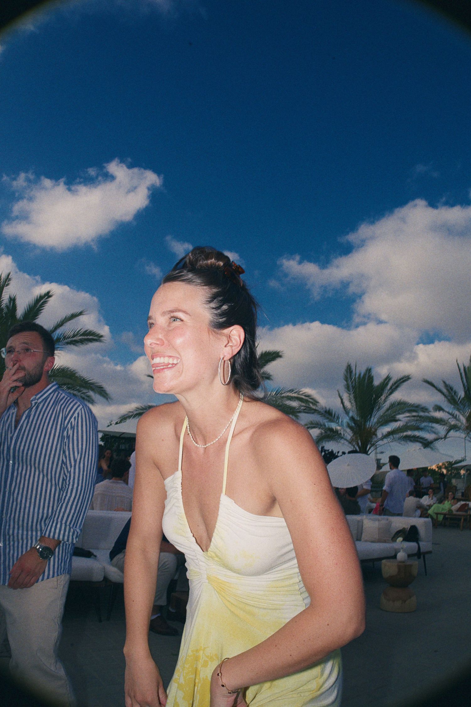 Young woman in yellow dress laughing outdoors at a beach party under blue sky with palm trees.