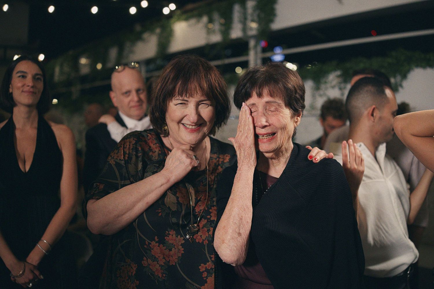 Two elderly women smile and laugh together on a lively wedding reception dance floor.