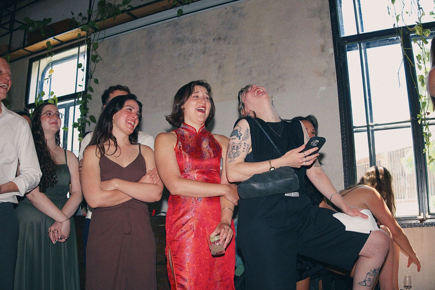 Wedding guests laughing and celebrating at a reception in an industrial venue with large windows.