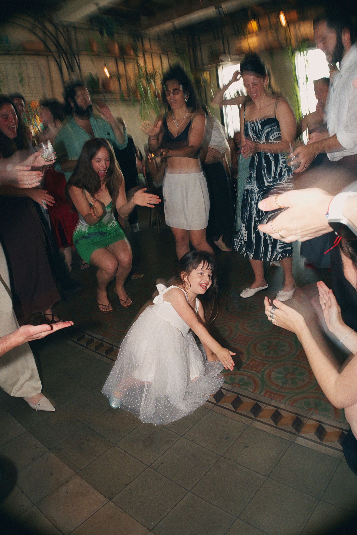 Bride in white tulle dress crouching on dance floor surrounded by dancing wedding guests at lively reception.