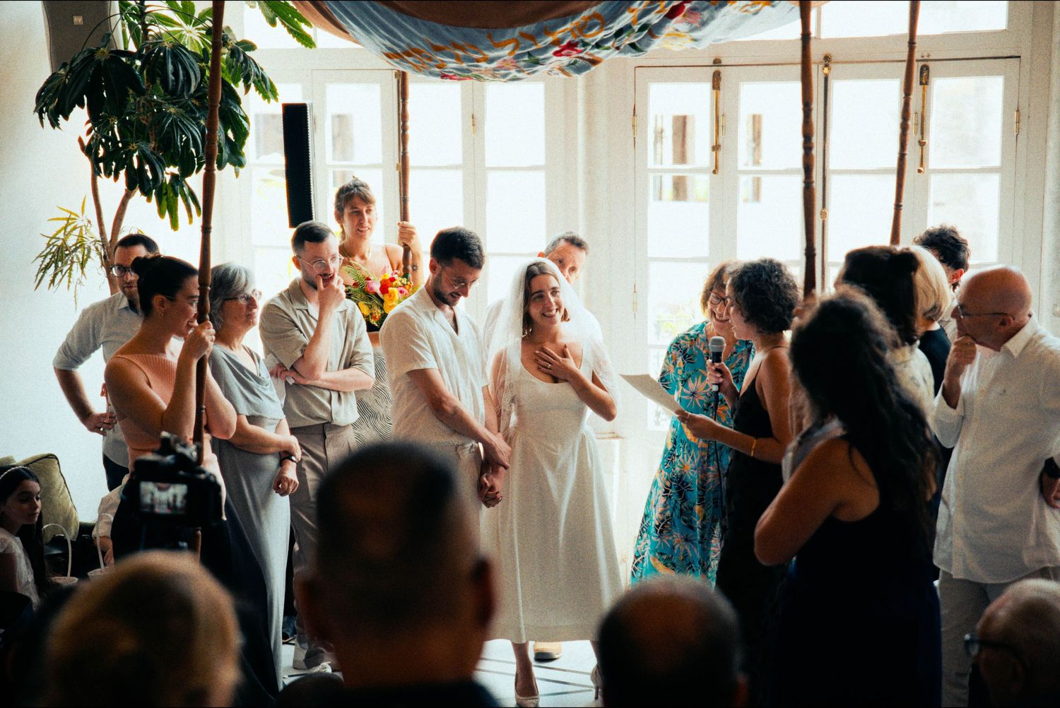 A group gathers for an intimate indoor wedding ceremony with natural lighting streaming through windows.