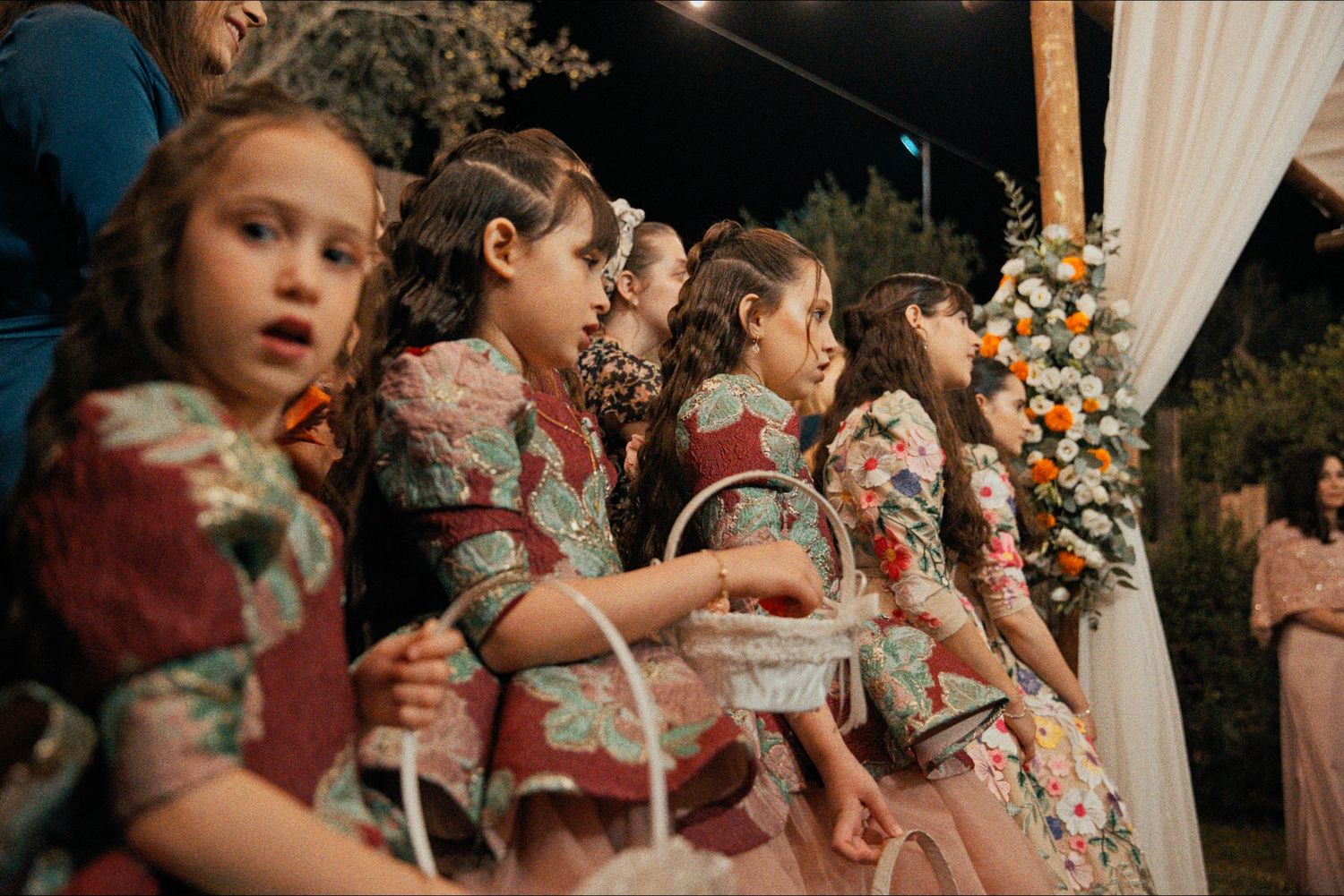 Row of flower girls in matching floral dresses at outdoor wedding ceremony.