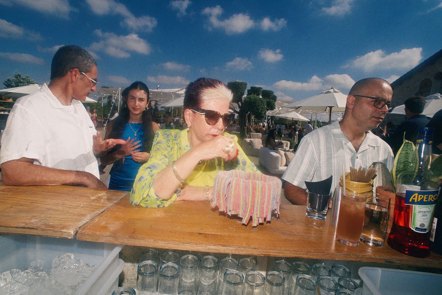 People socializing at an outdoor bar with drinks and Aperol bottles on a sunny day.