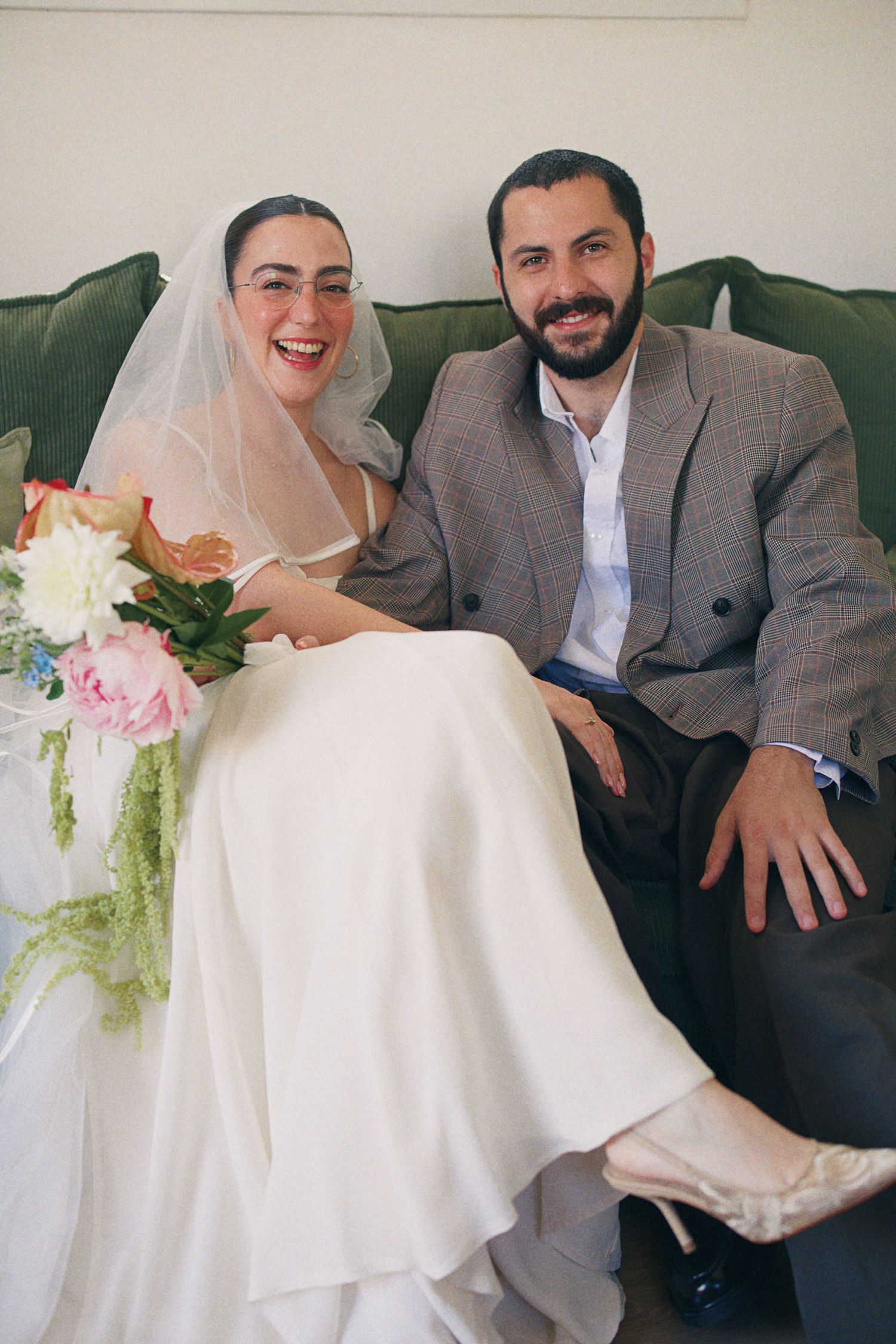 Smiling bride in white gown and veil holding floral bouquet sits with bearded groom in gray suit on green sofa.