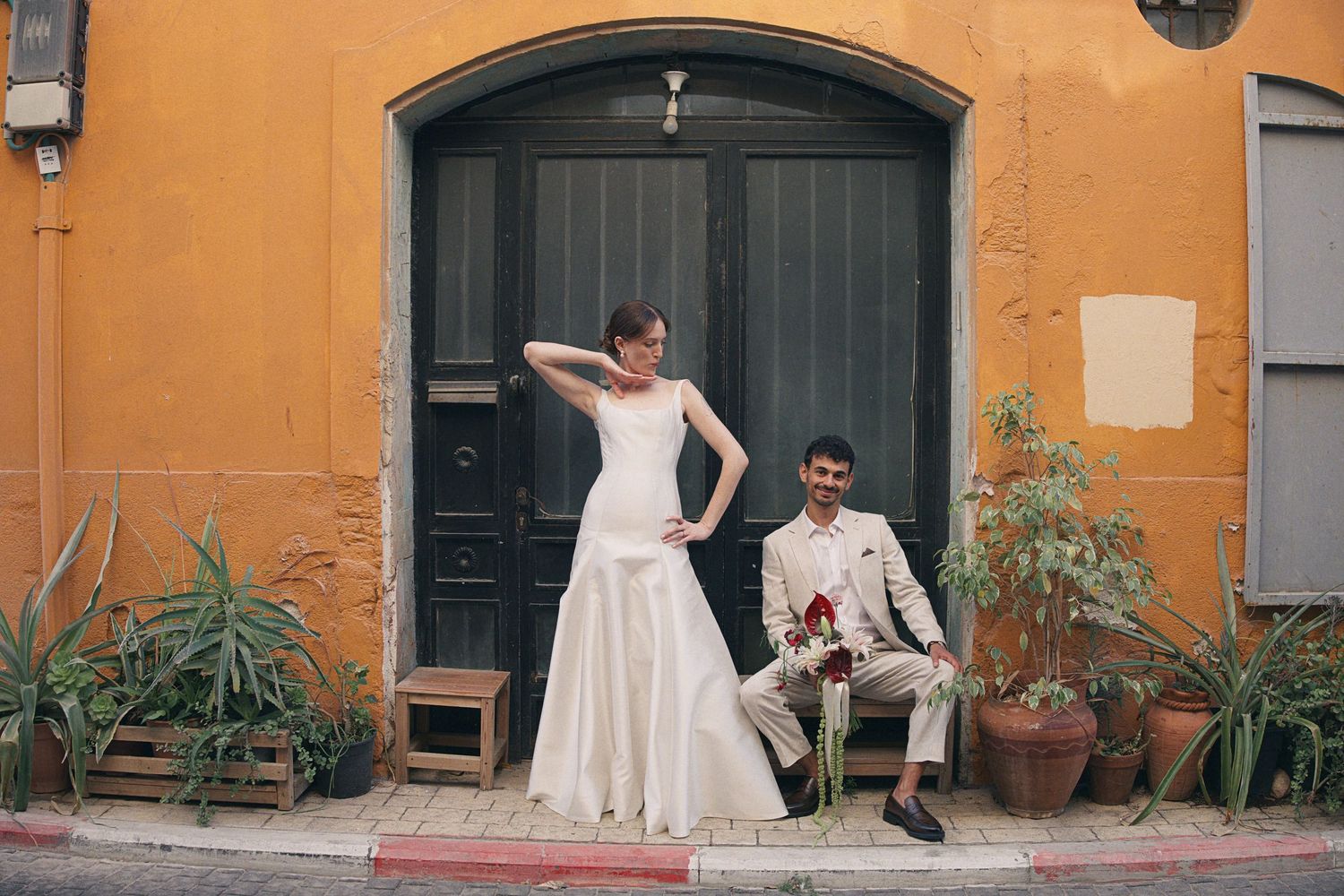 Bride in white mermaid gown and groom in tan suit pose before yellow building with green door.