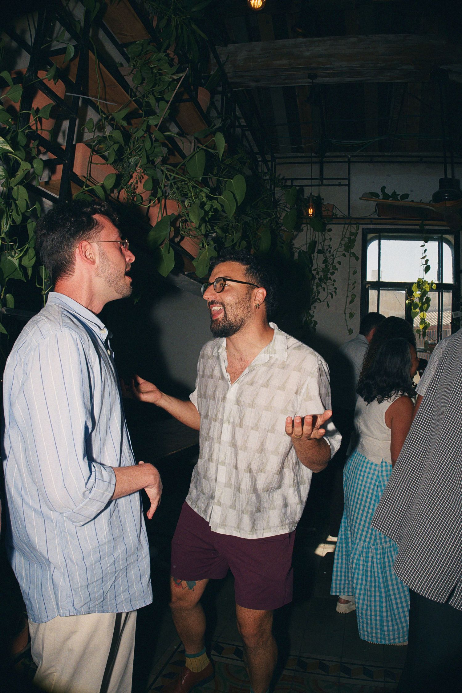 Two men in casual shirts laugh together at an indoor wedding reception.