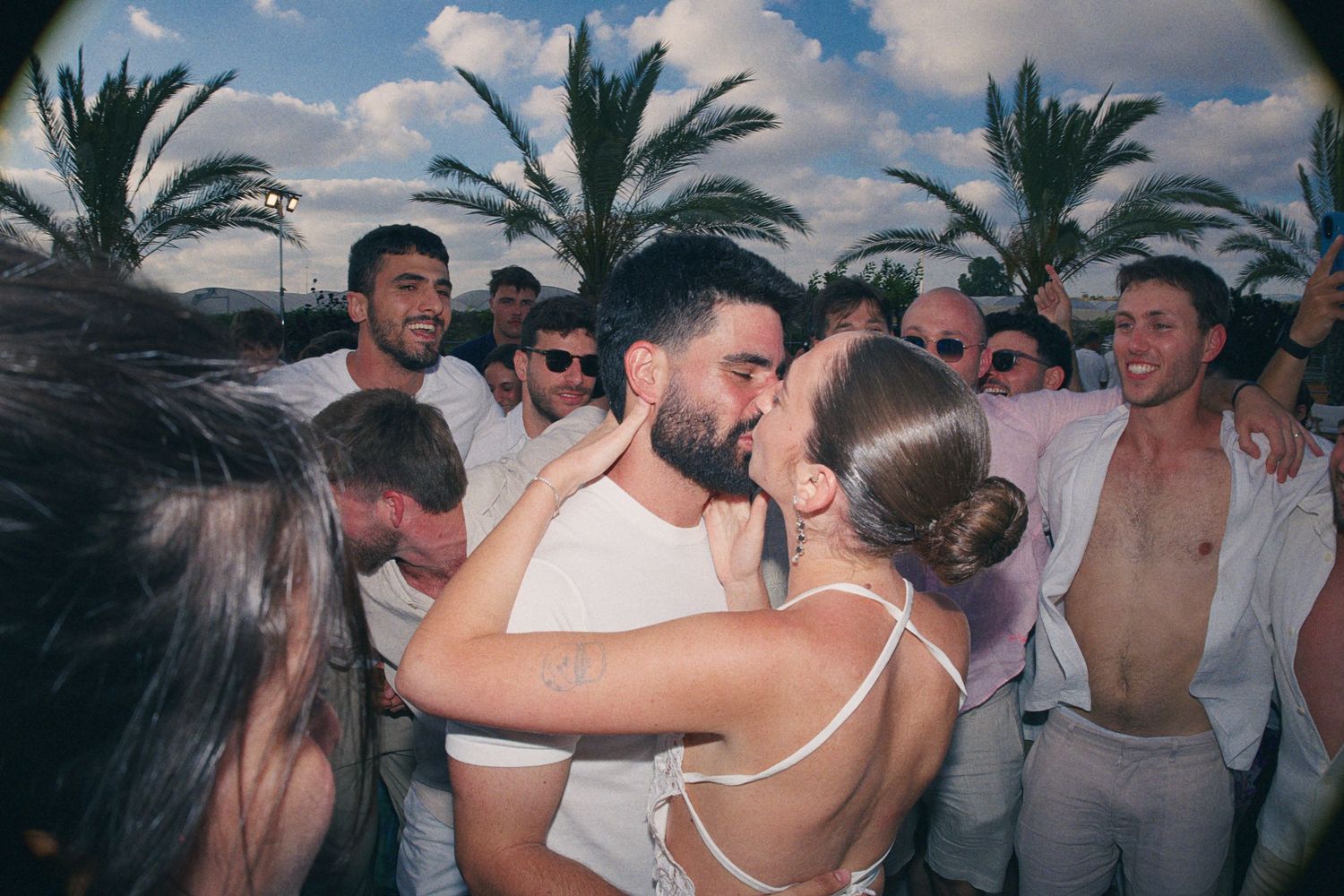 Couple shares romantic kiss surrounded by guests dressed in white at lively outdoor wedding reception.