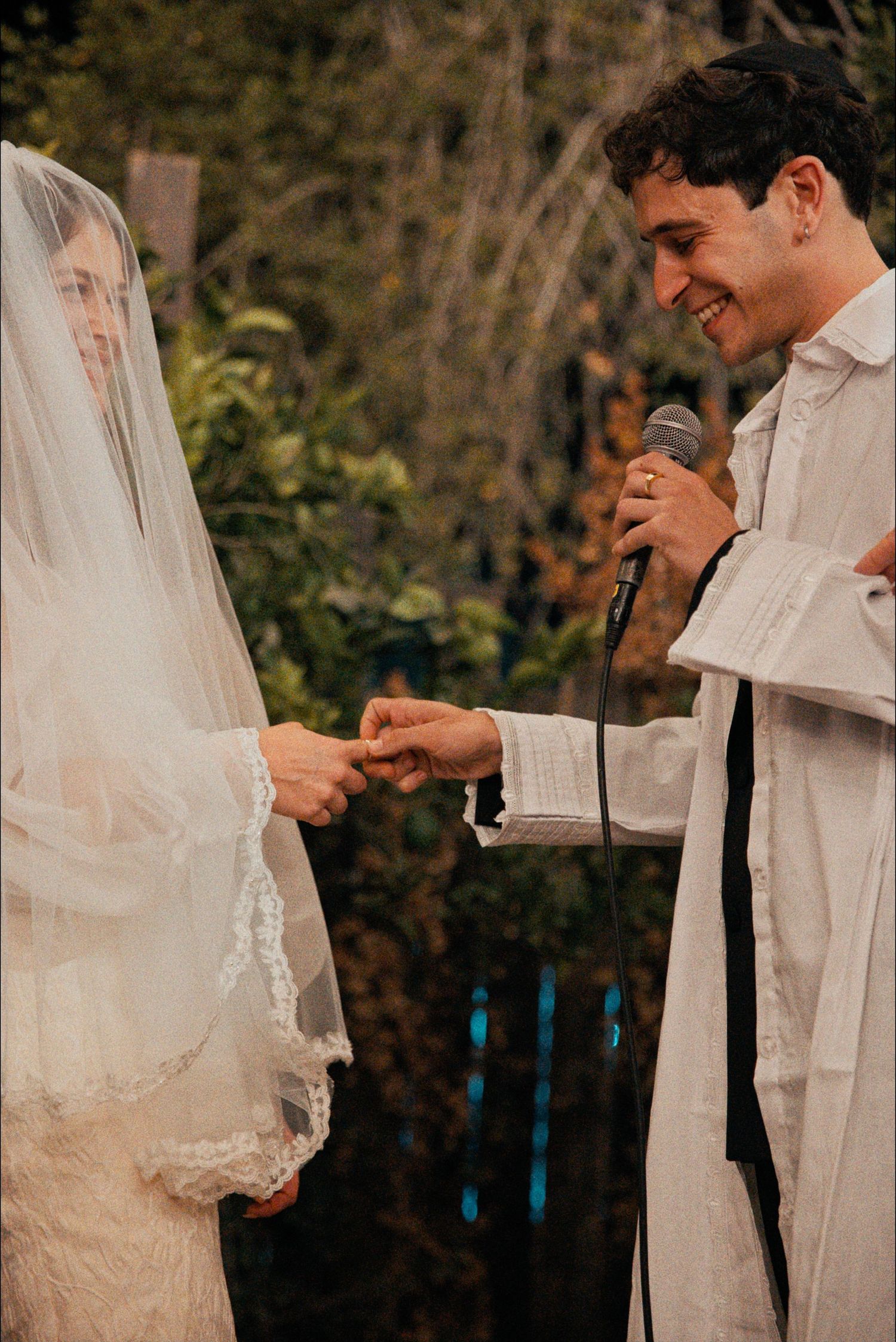 Exchange of rings during outdoor wedding ceremony under wooden arch with white draping.