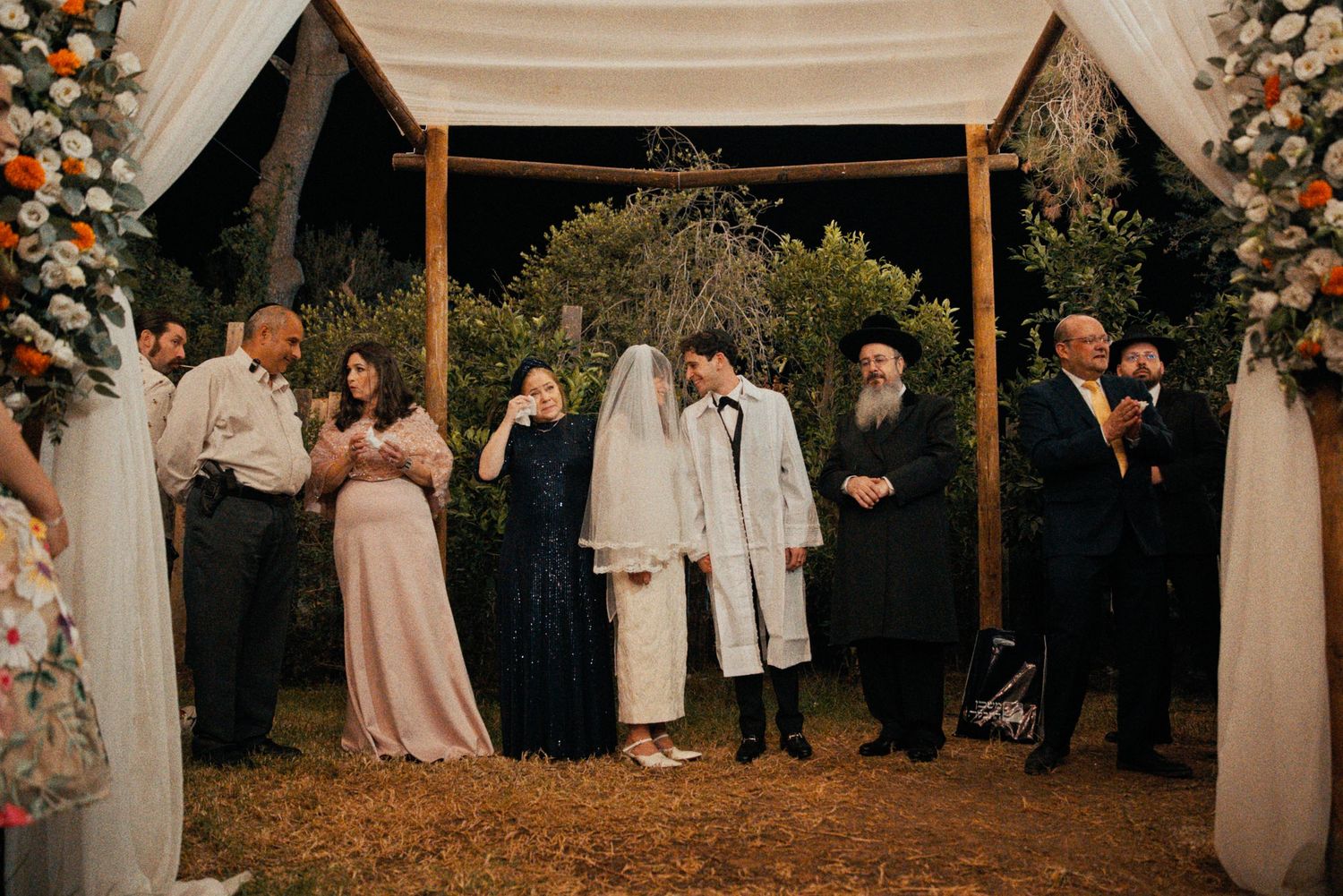 Wedding ceremony under a rustic wooden arch decorated with white drapes and floral arrangements.