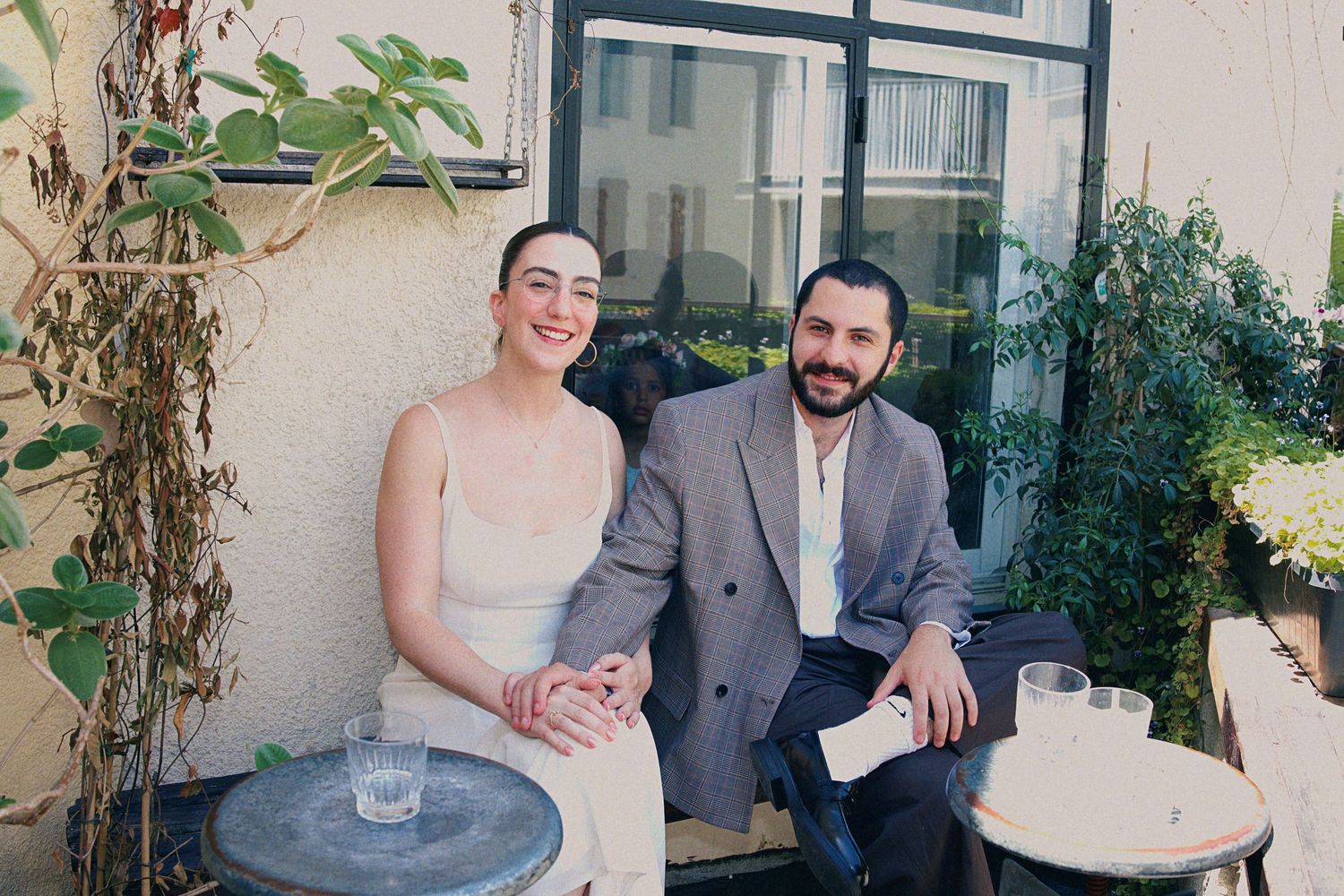 Couple smiling together on outdoor patio with plants, woman in white dress and man in gray blazer.