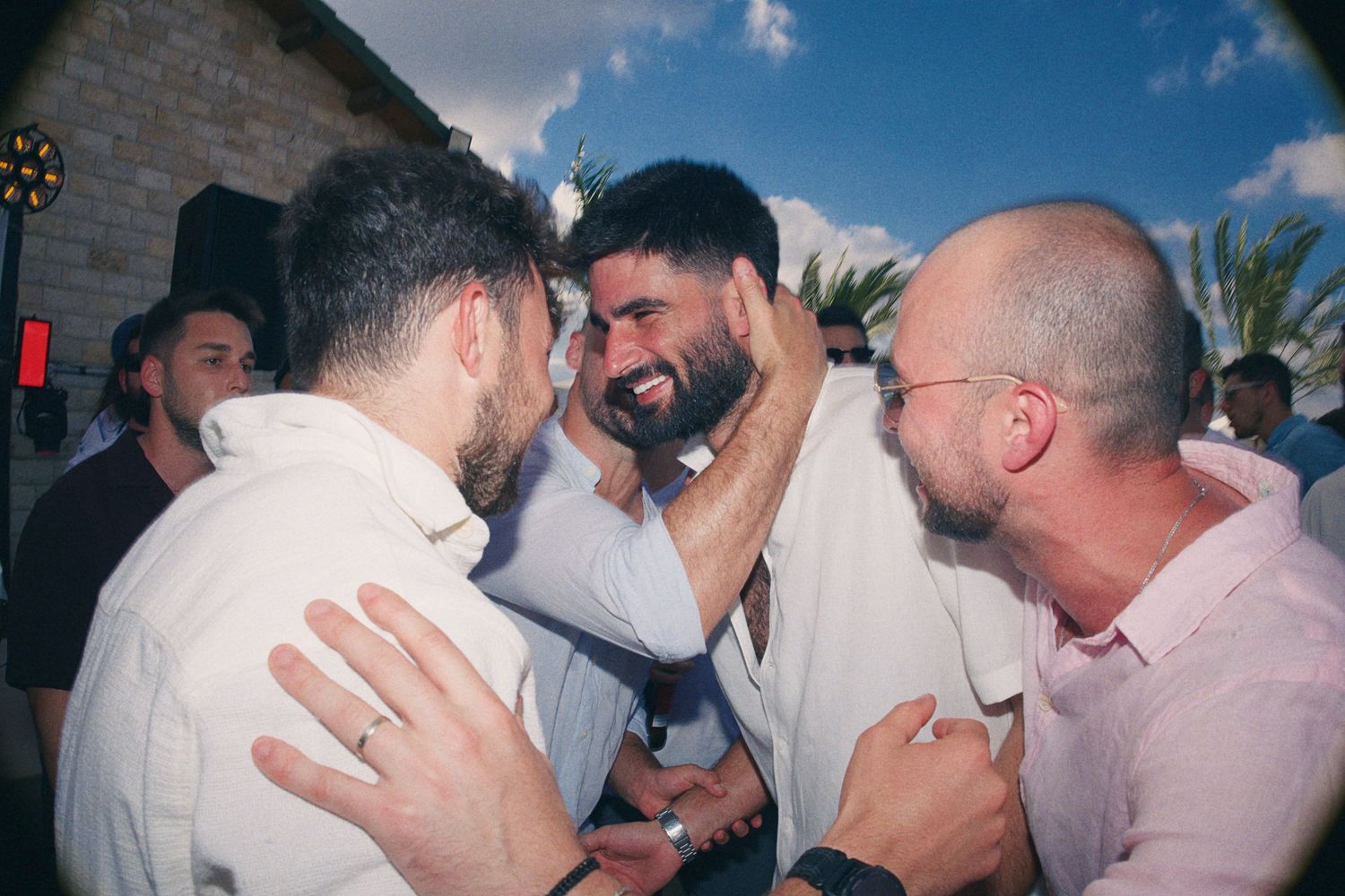 Three bearded men in white shirts share an emotional embrace at an outdoor celebration event.