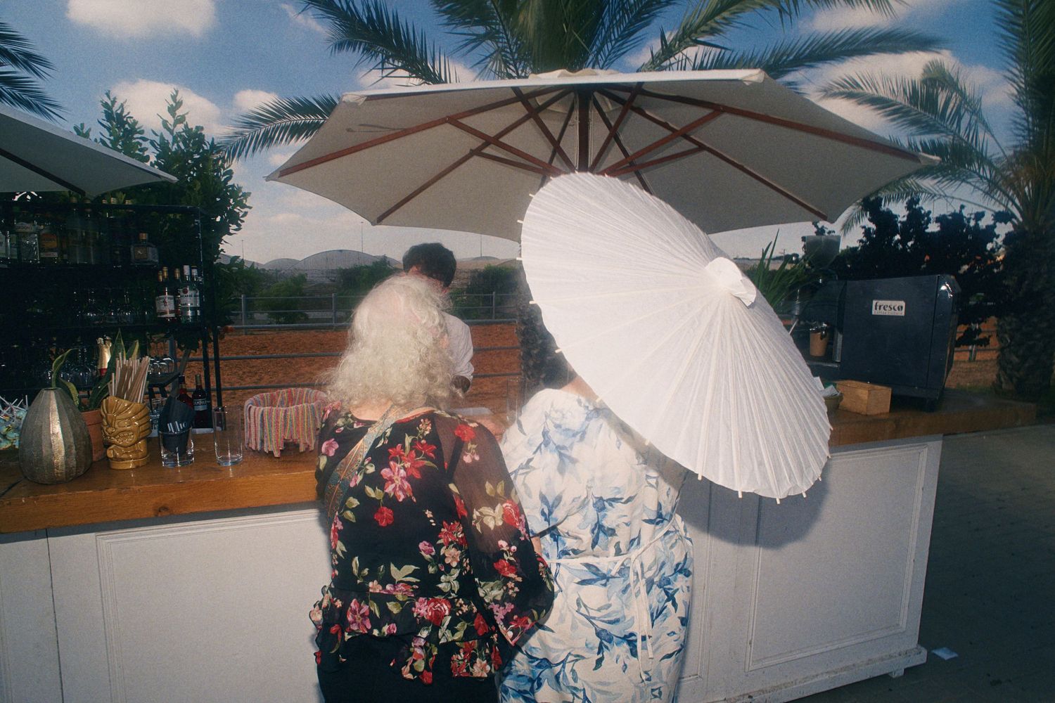 Two women holding white parasols stand at an outdoor bar with a large patio umbrella and palm trees.