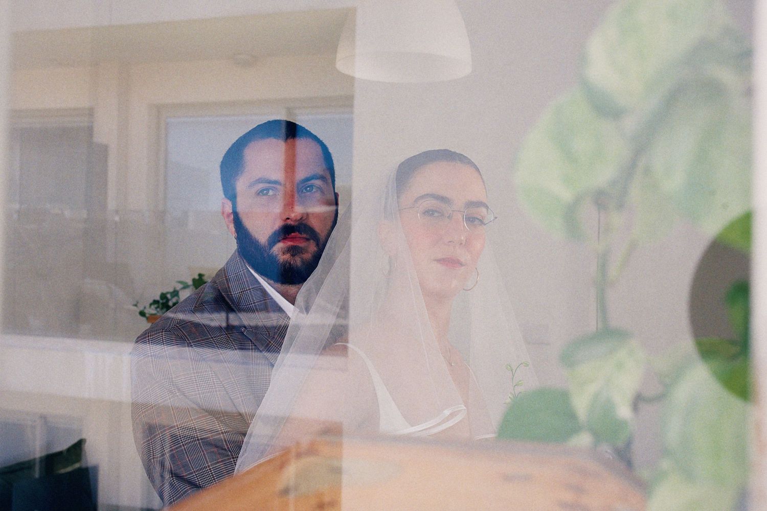 Bride and groom reflected in glass window, wearing wedding attire with veil and gray suit.