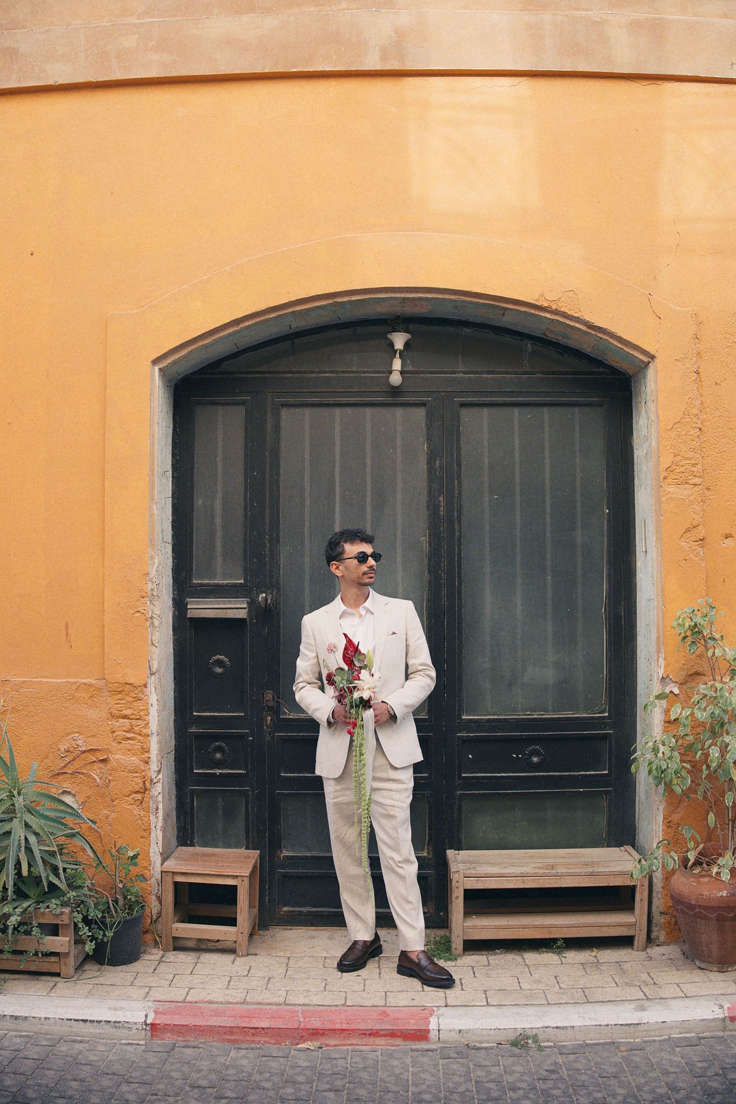 Stylish man in beige suit holding red roses standing before arched green door on yellow wall.