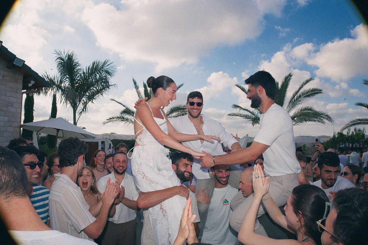Bride lifted by groom above cheering crowd at outdoor wedding celebration with palm trees in background.