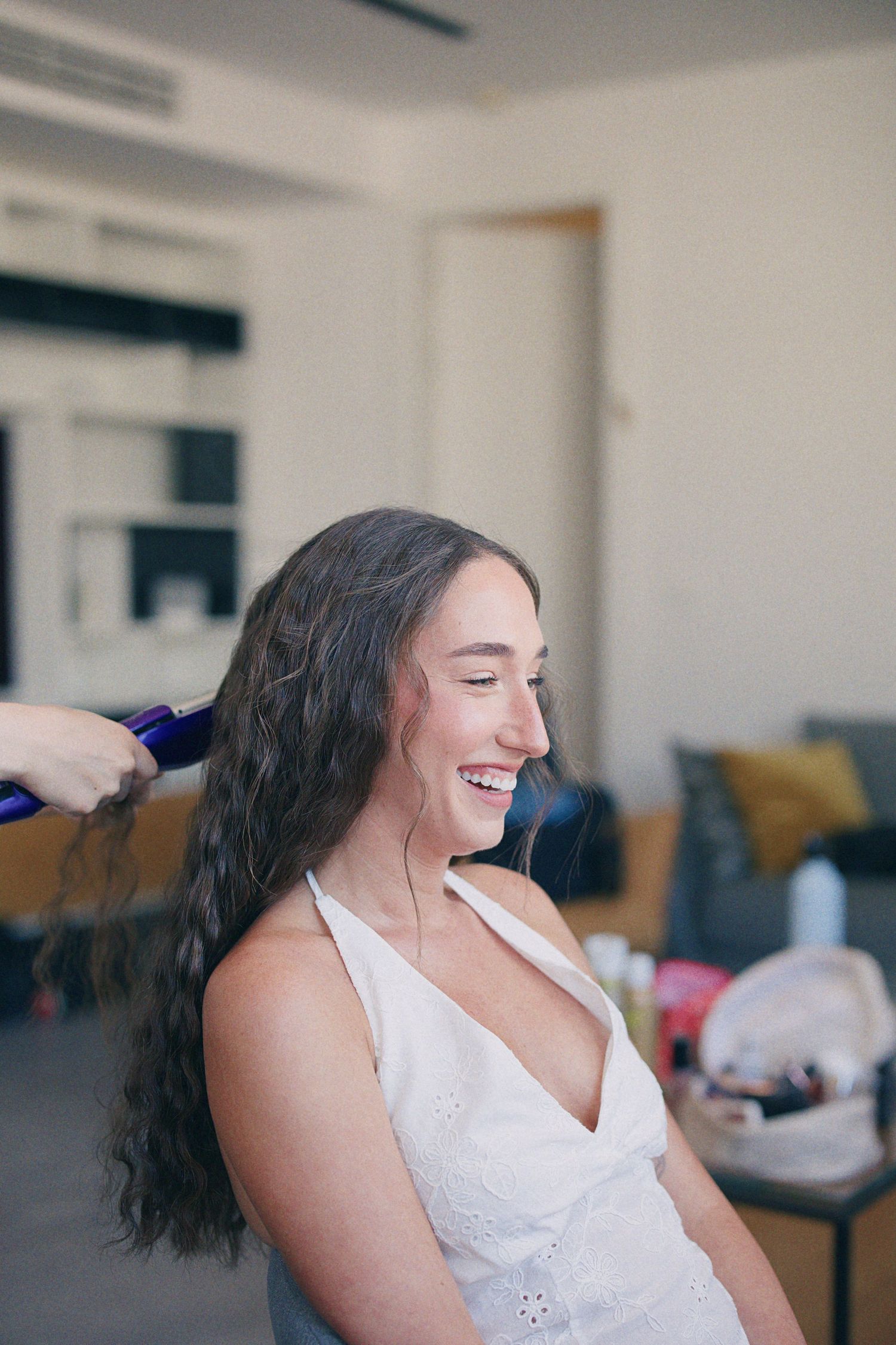 Smiling bride getting her long dark hair styled with a curling iron before her wedding ceremony.