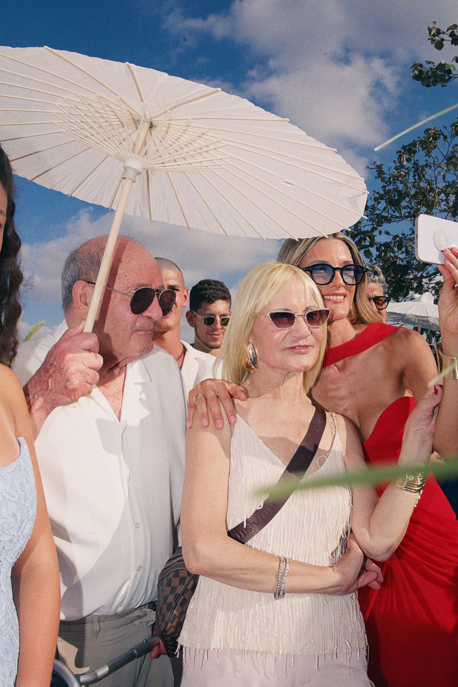 Wedding guests gather outdoors under a white parasol on a sunny day with blue skies.