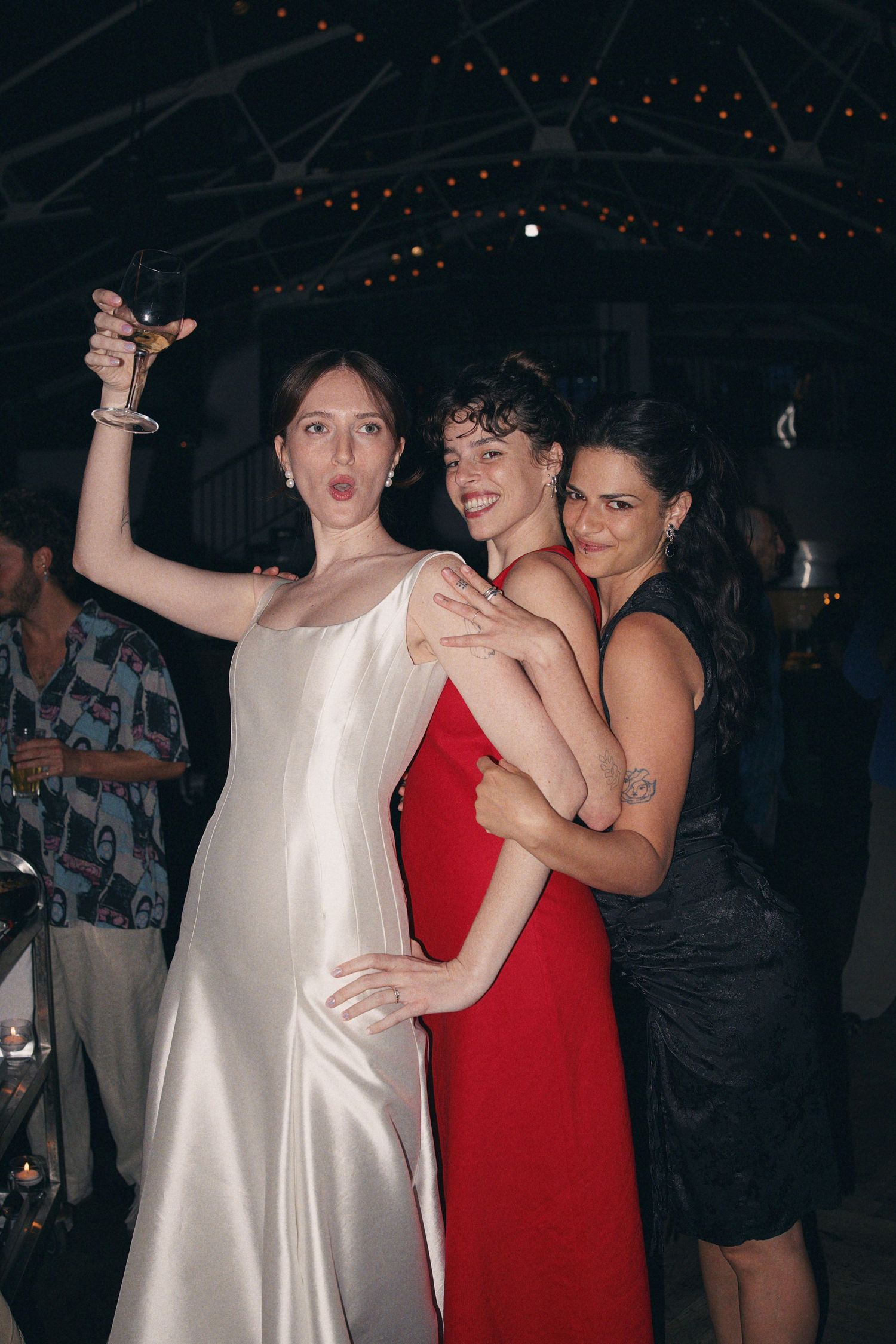 Bride in white satin gown raises champagne glass while two friends in red and black dresses embrace her.