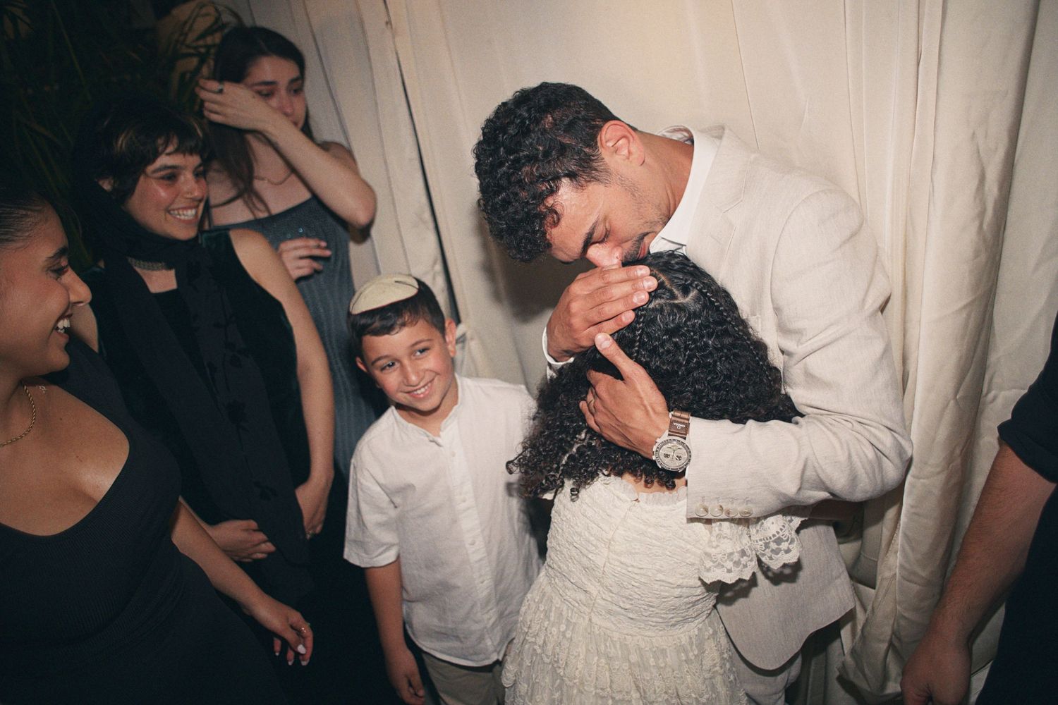 Man in white suit kissing a woman's head at a celebration while children and guests watch and smile nearby.