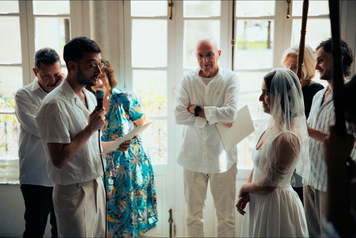 Wedding guests stand together during a ceremony in a bright sunlit room with large windows.