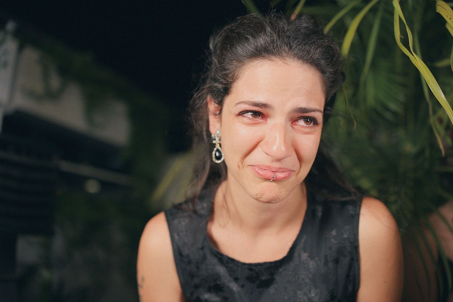 Emotional woman with teary eyes wearing drop earrings and black dress at an outdoor evening event.