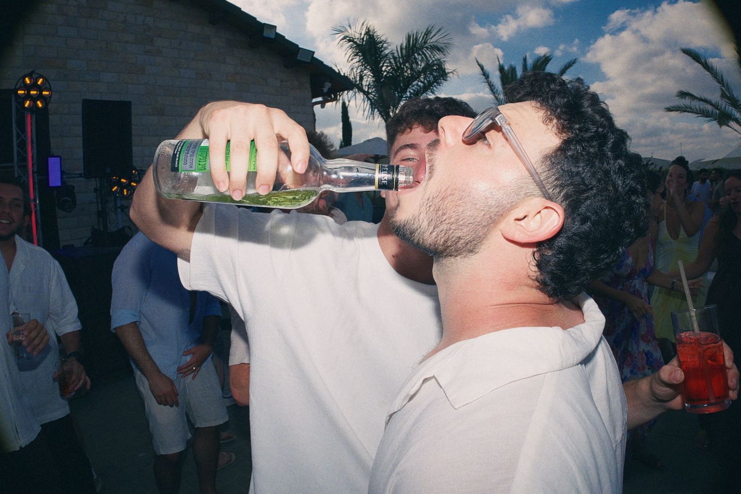 Man in white shirt drinking from bottle at outdoor evening party with palm trees in background.