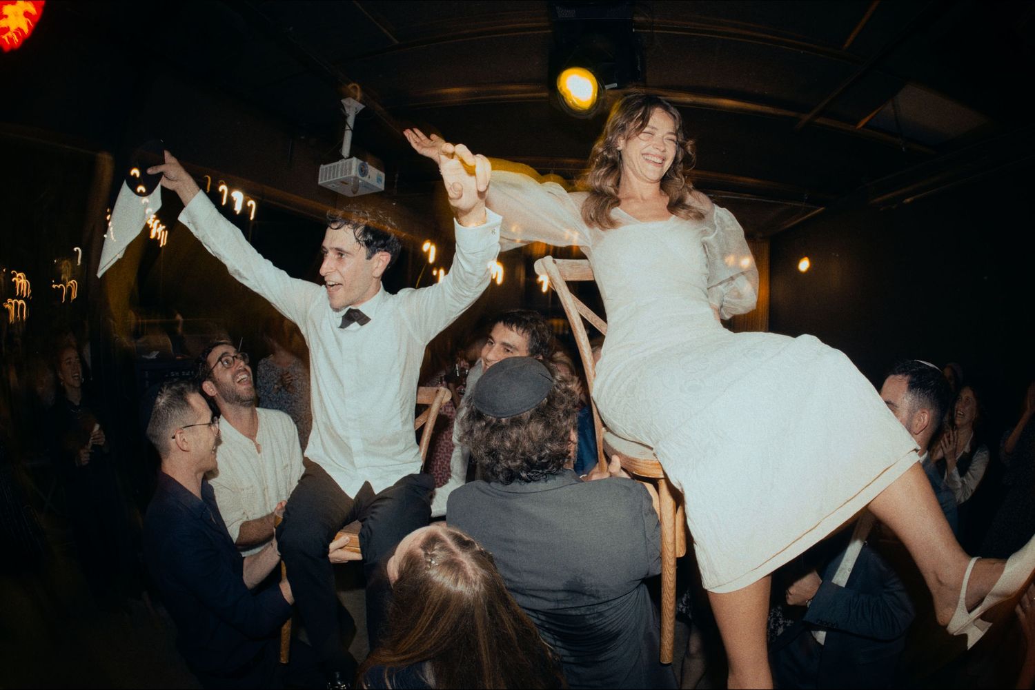 Wedding guests celebrate on the dance floor with energetic dancing and lifting at an indoor reception venue.