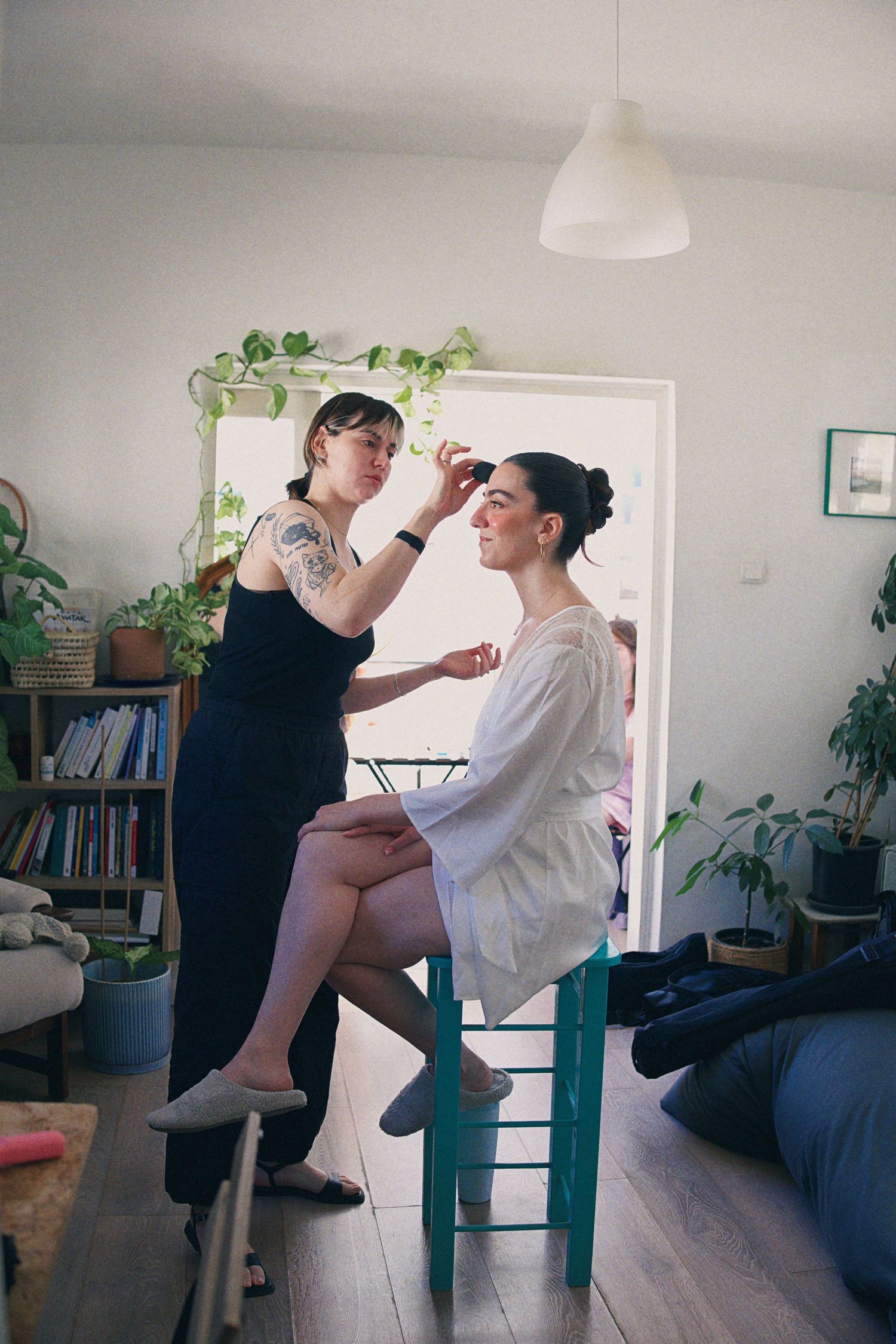 Makeup artist applying makeup to a bride in a white robe seated on a teal chair in a plant-filled apartment.