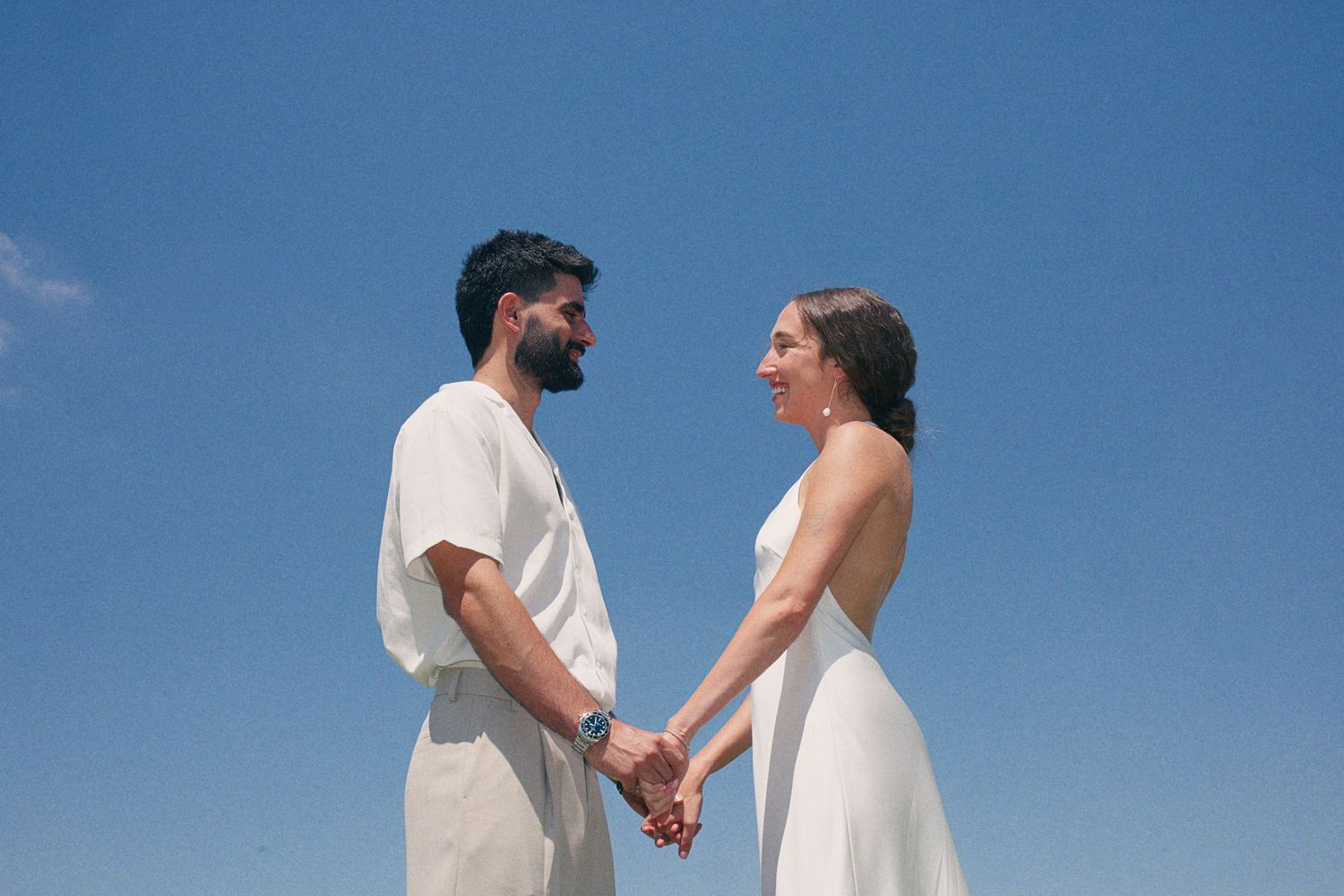 Couple in white wedding attire hold hands facing each other against a bright blue sky.