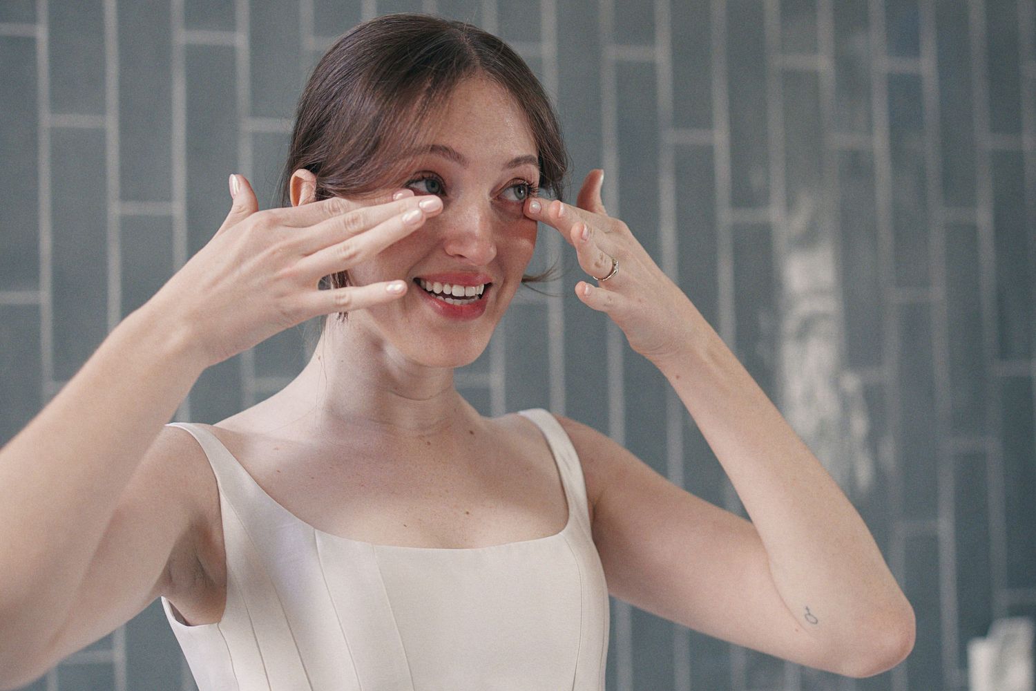 Young woman smiling while applying skincare product to her face in a modern bathroom.