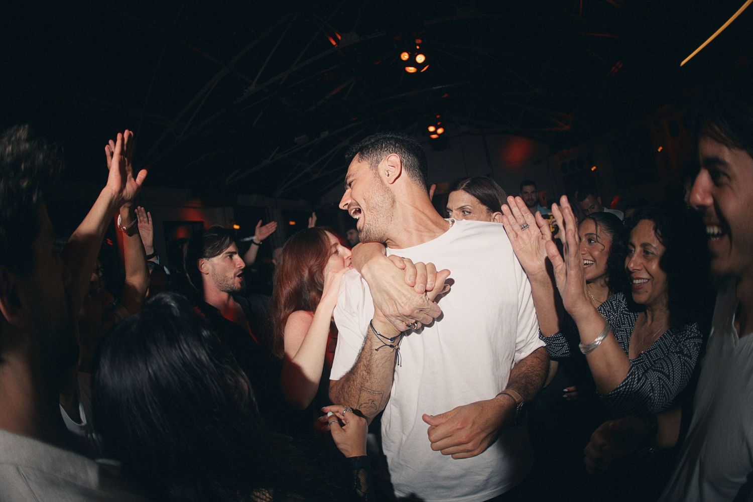 Crowd of people dancing and celebrating at a nightclub party, with a man in a white t-shirt at the center.
