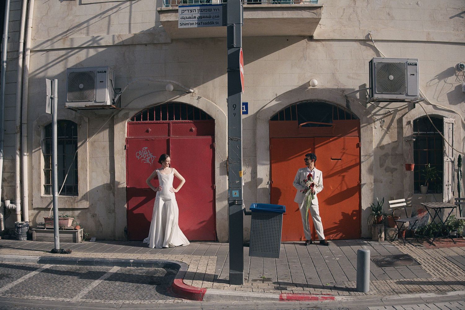 Bride and groom pose separately by red doors on a sunlit urban street corner.