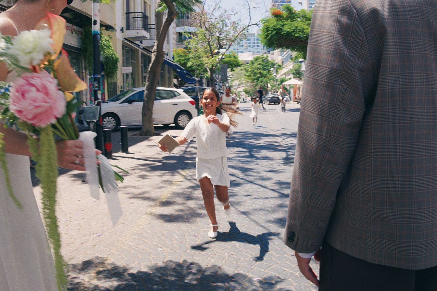 Excited flower girl runs toward wedding couple on sunny urban street during outdoor ceremony.