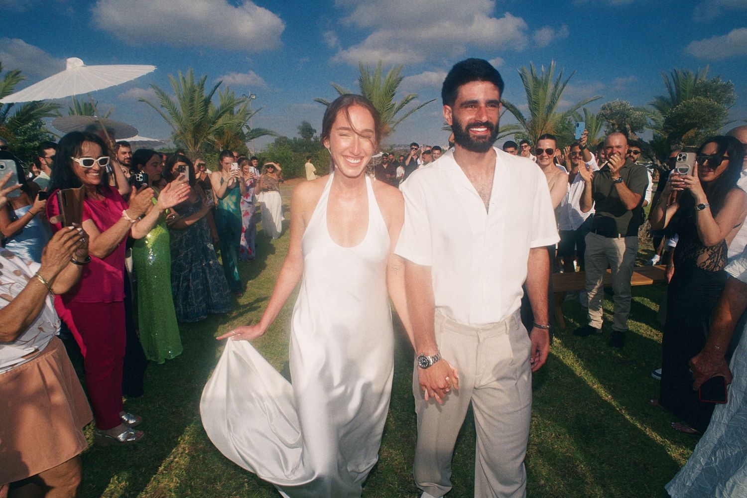 Couple in white wedding attire walks through cheering guests outdoors with palm trees at a sunny destination wedding.