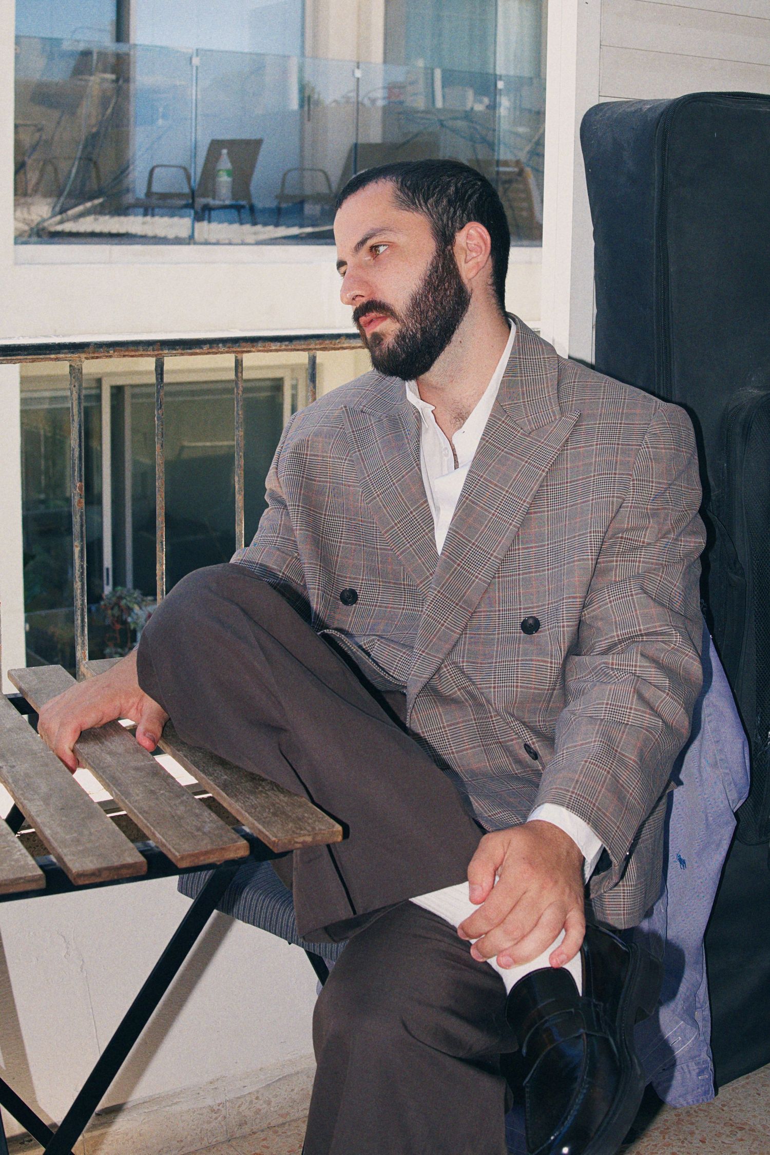 Bearded man in plaid blazer sits on wooden bench on balcony looking away.
