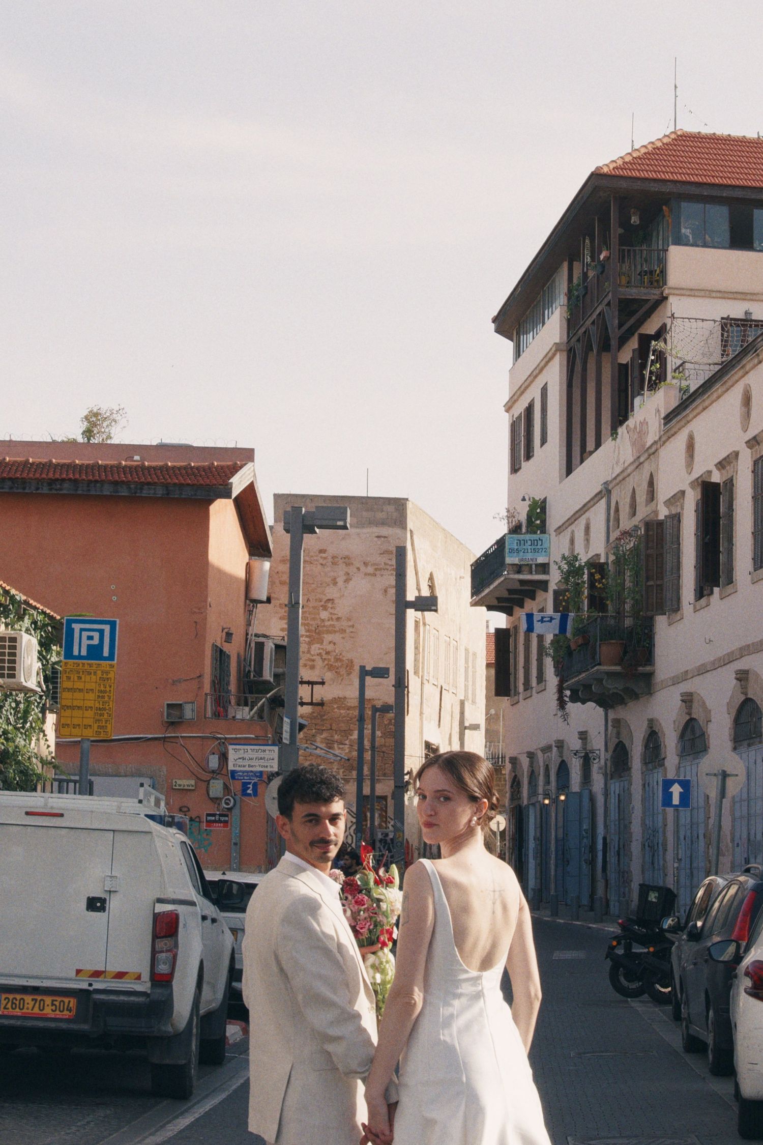 Bride and groom holding hands on a city street, bride in white gown, groom in beige suit.