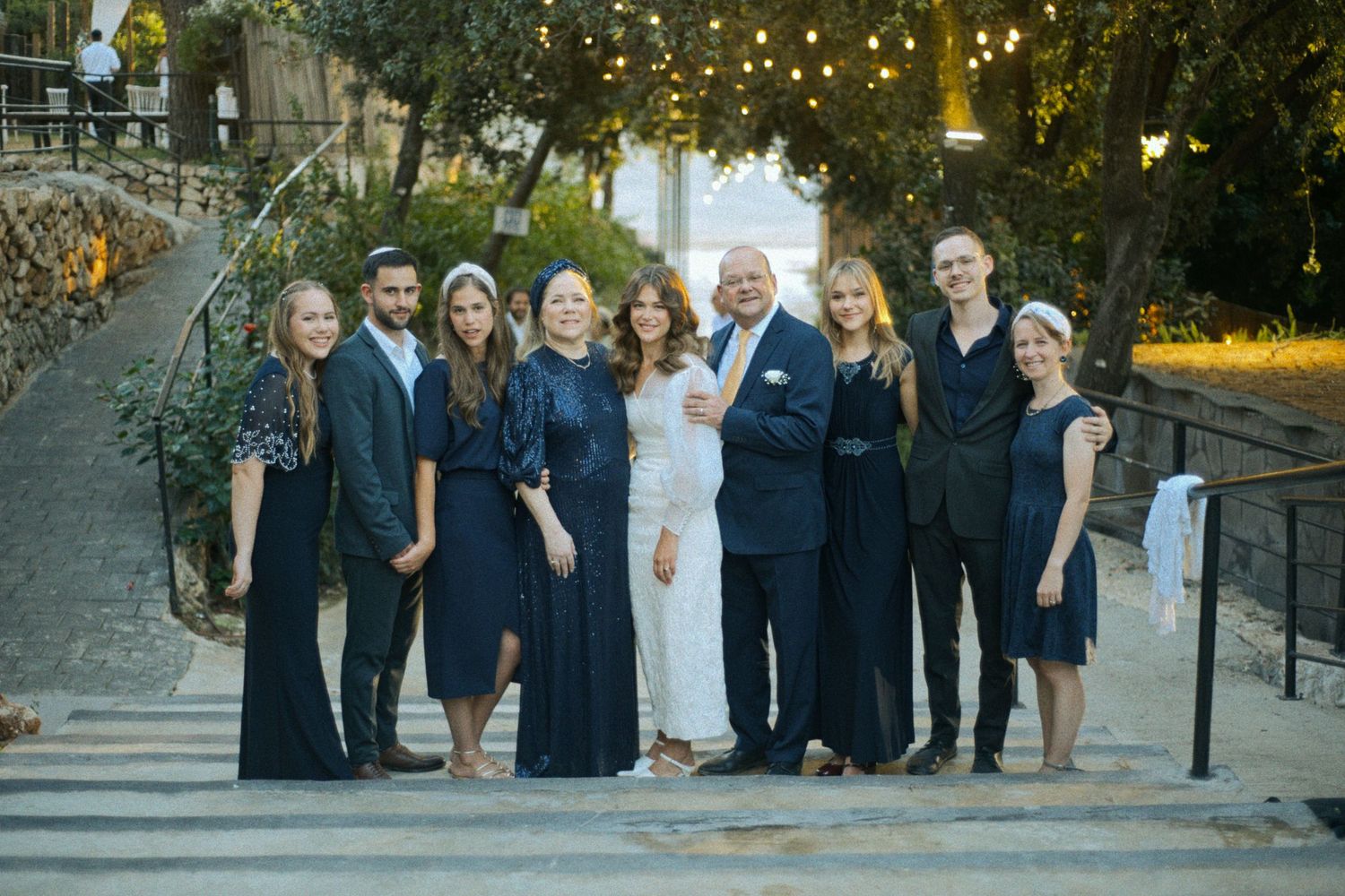 Group photo of wedding attendees in navy and white attire standing together under string lights.