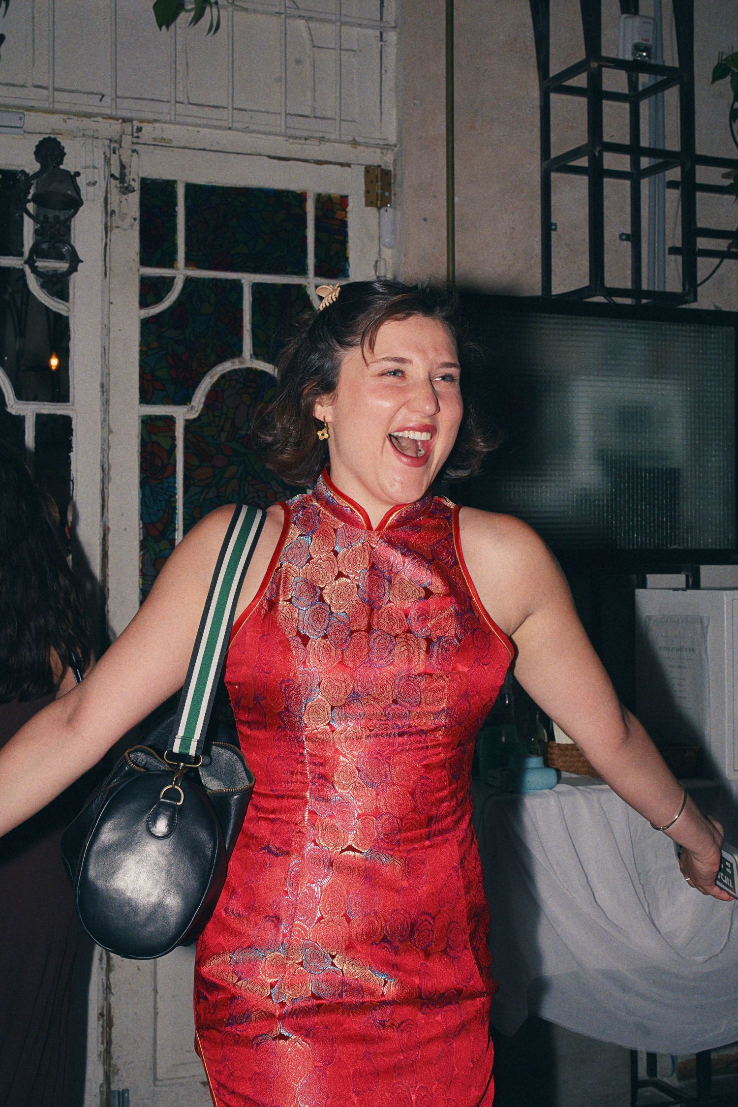 Woman in red floral cheongsam dress laughing joyfully at a party, carrying a black leather handbag.